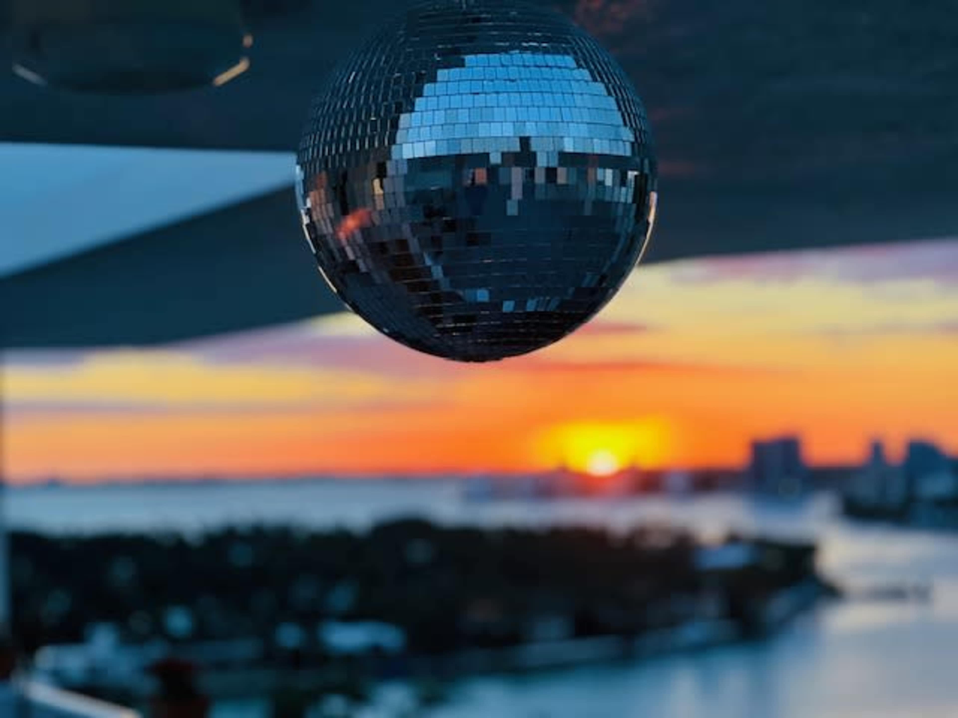A disco ball hangs in the foreground as the sun sets over a waterfront city skyline.