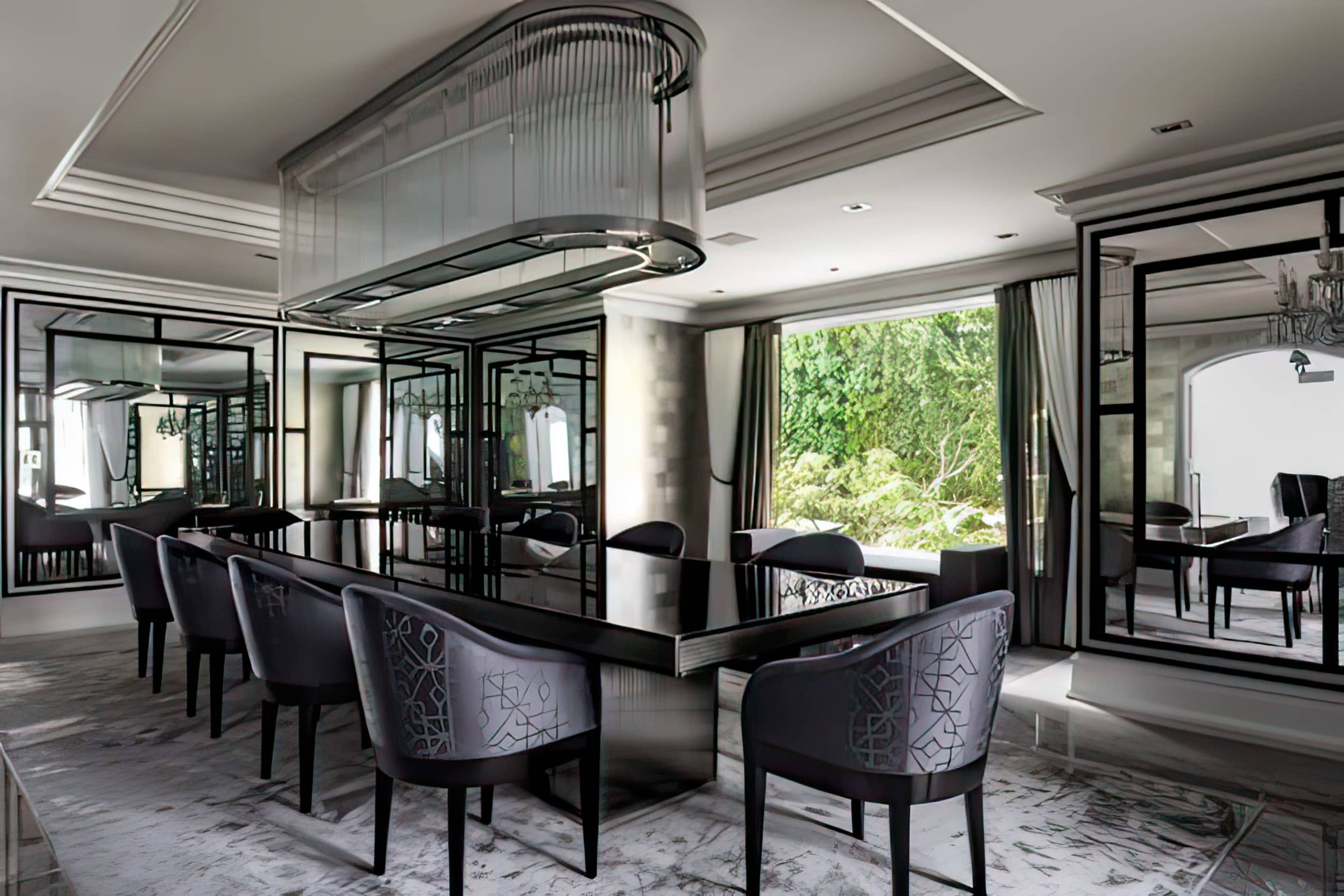 A modern dining room features a long black table surrounded by upholstered chairs, with large mirrors and windows reflecting the greenery outside.