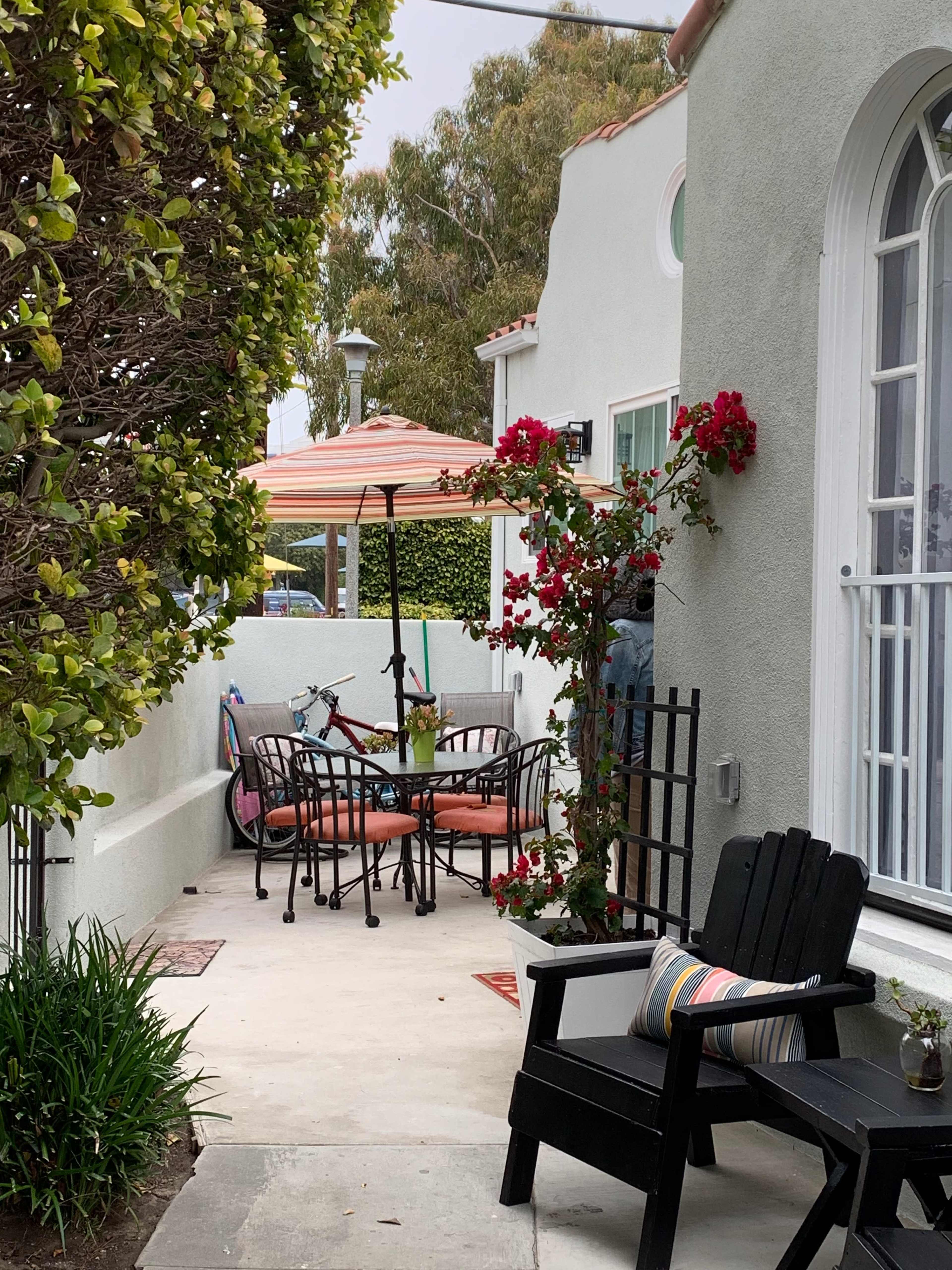 A narrow pathway leads to a patio with a table and chairs, surrounded by greenery and blooming flowers.