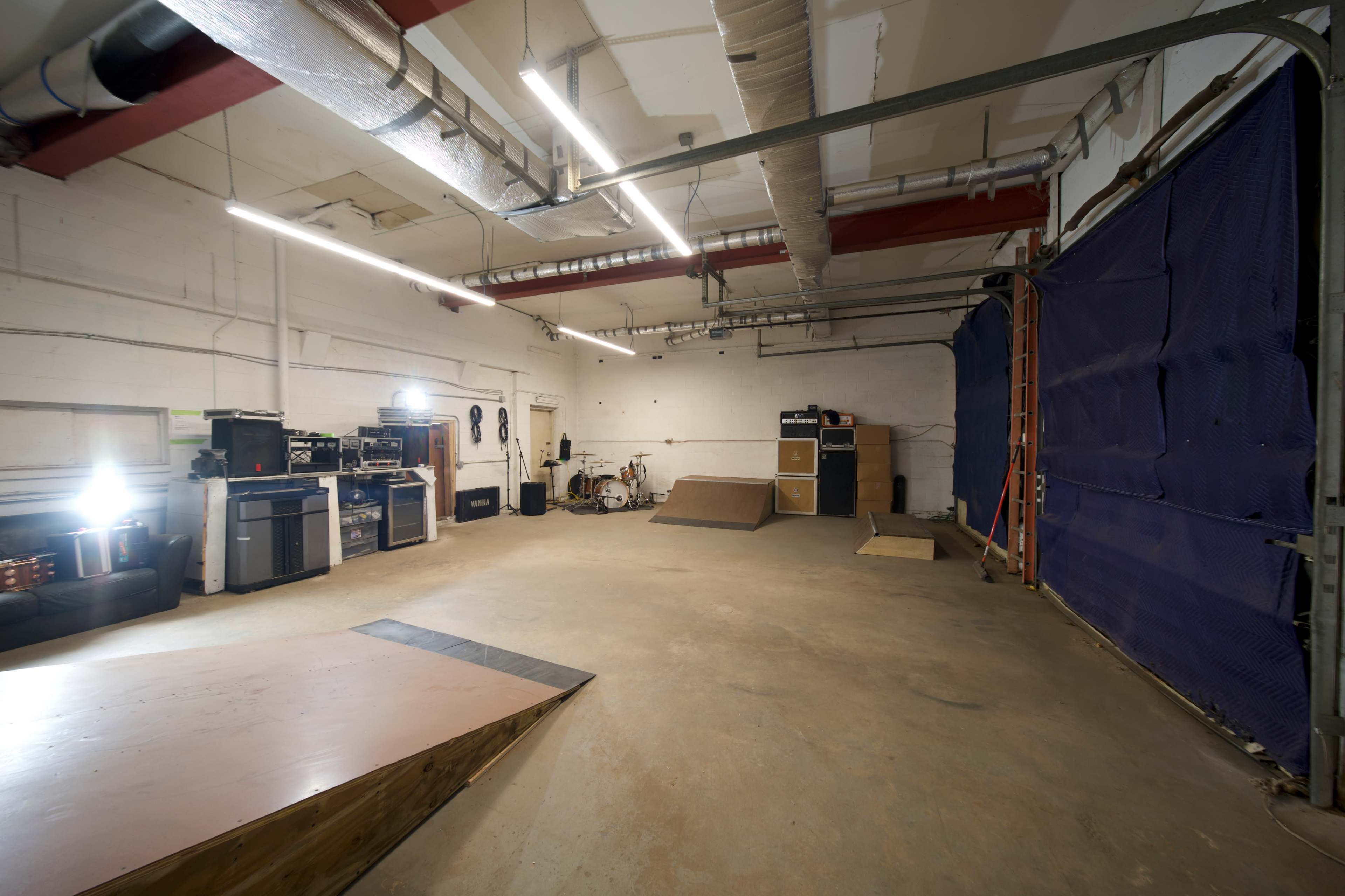 An empty rehearsal space with musical equipment, a small ramp, and a blue curtain at one end.