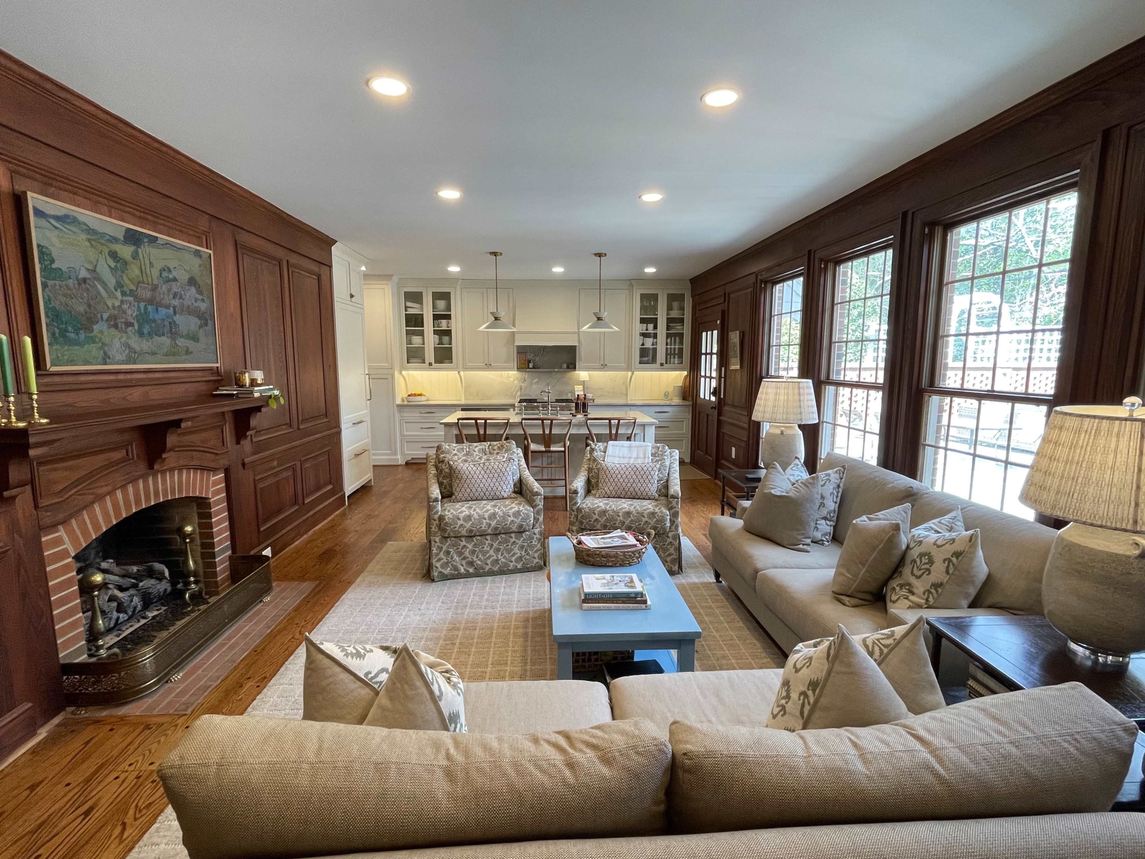 The image shows a cozy living room with two upholstered sofas and armchairs, a coffee table, a fireplace, and an open kitchen in the background.