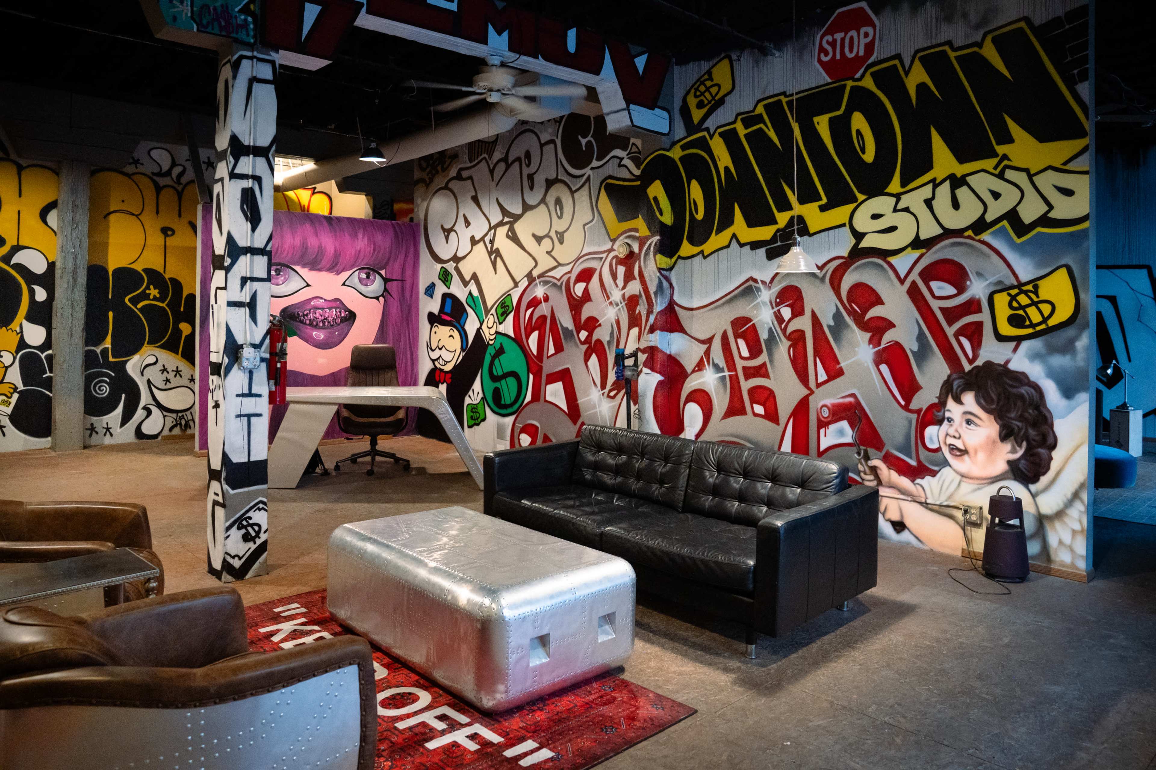 The scene features a vibrant studio space with graffiti-covered walls, a modern black couch, and a desk area, creating an artistic urban environment.