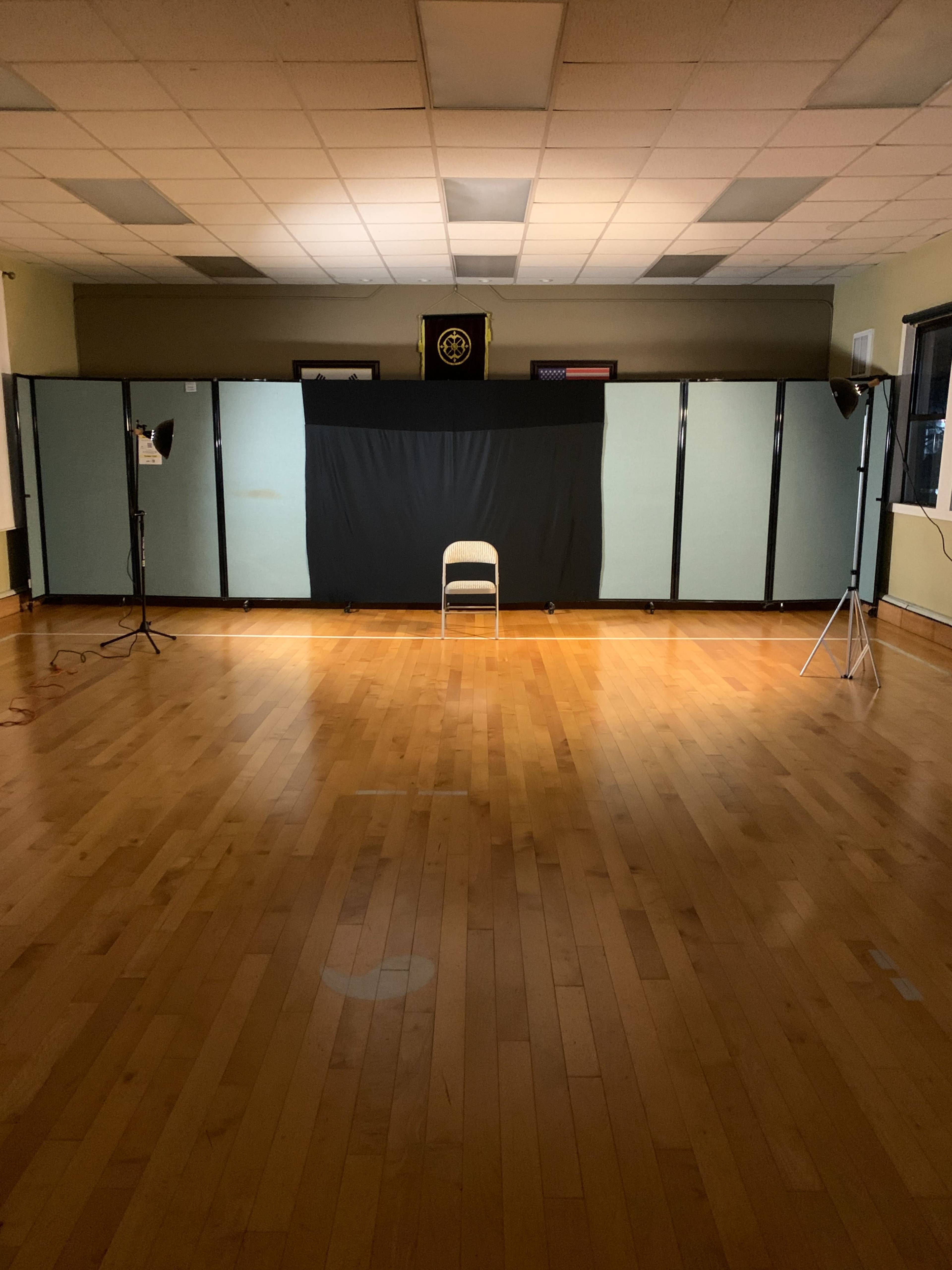 The image shows an empty room with a wooden floor, featuring a single chair in front of a black backdrop, illuminated by two lights on either side.
