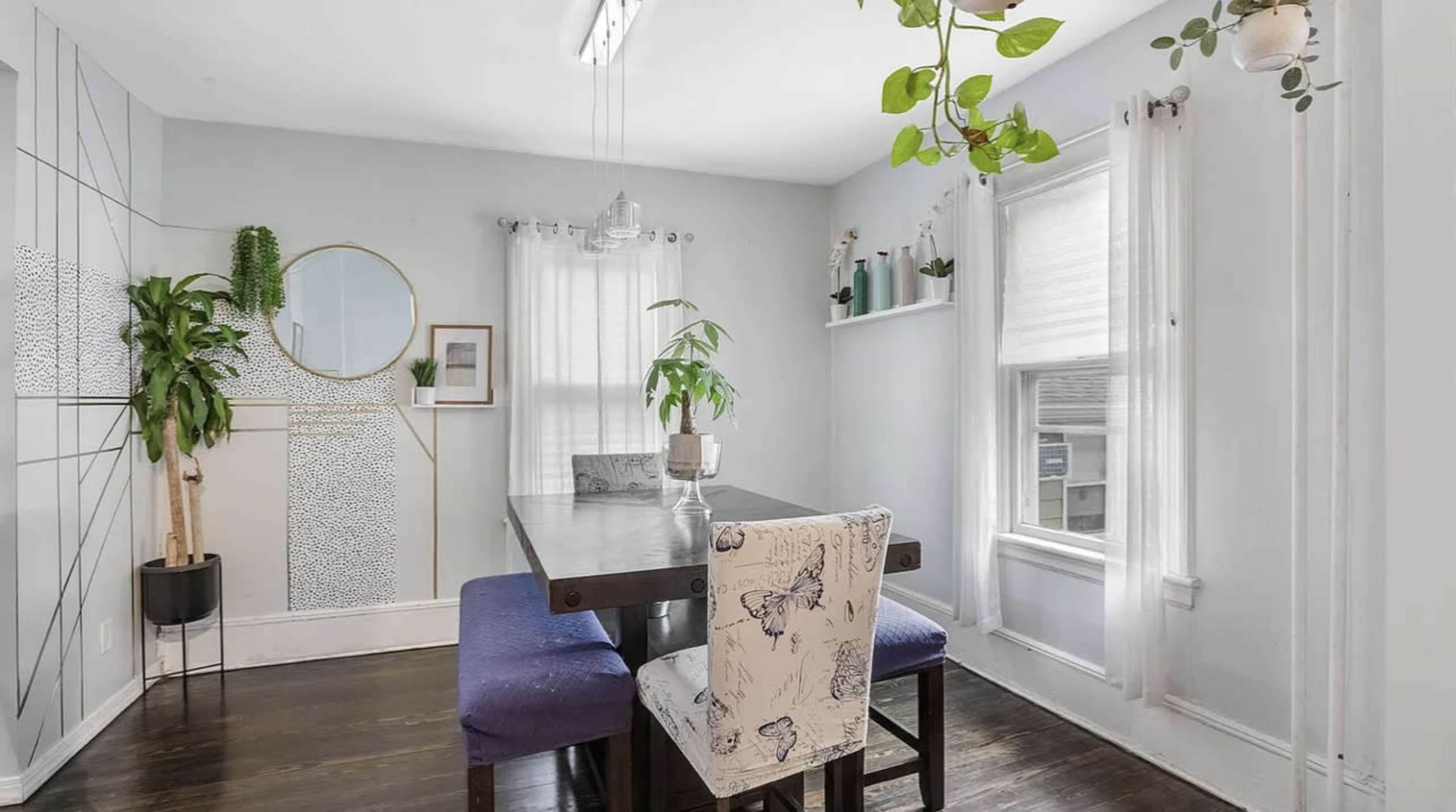 A dining area features a table with four chairs, plants decorating the space, and a large mirror on the wall.