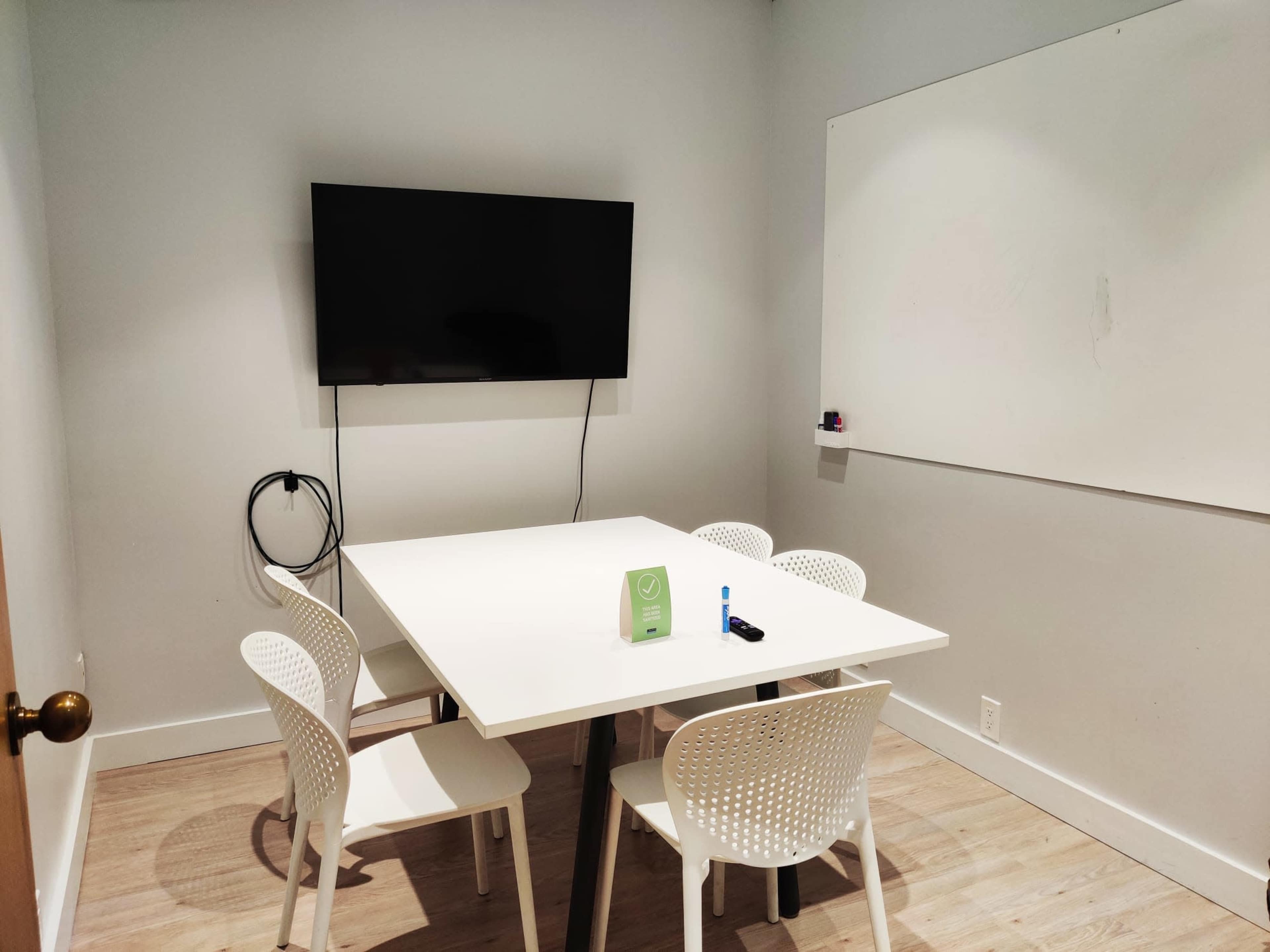 A small meeting room features a white table with four chairs, a wall-mounted television, and a whiteboard.