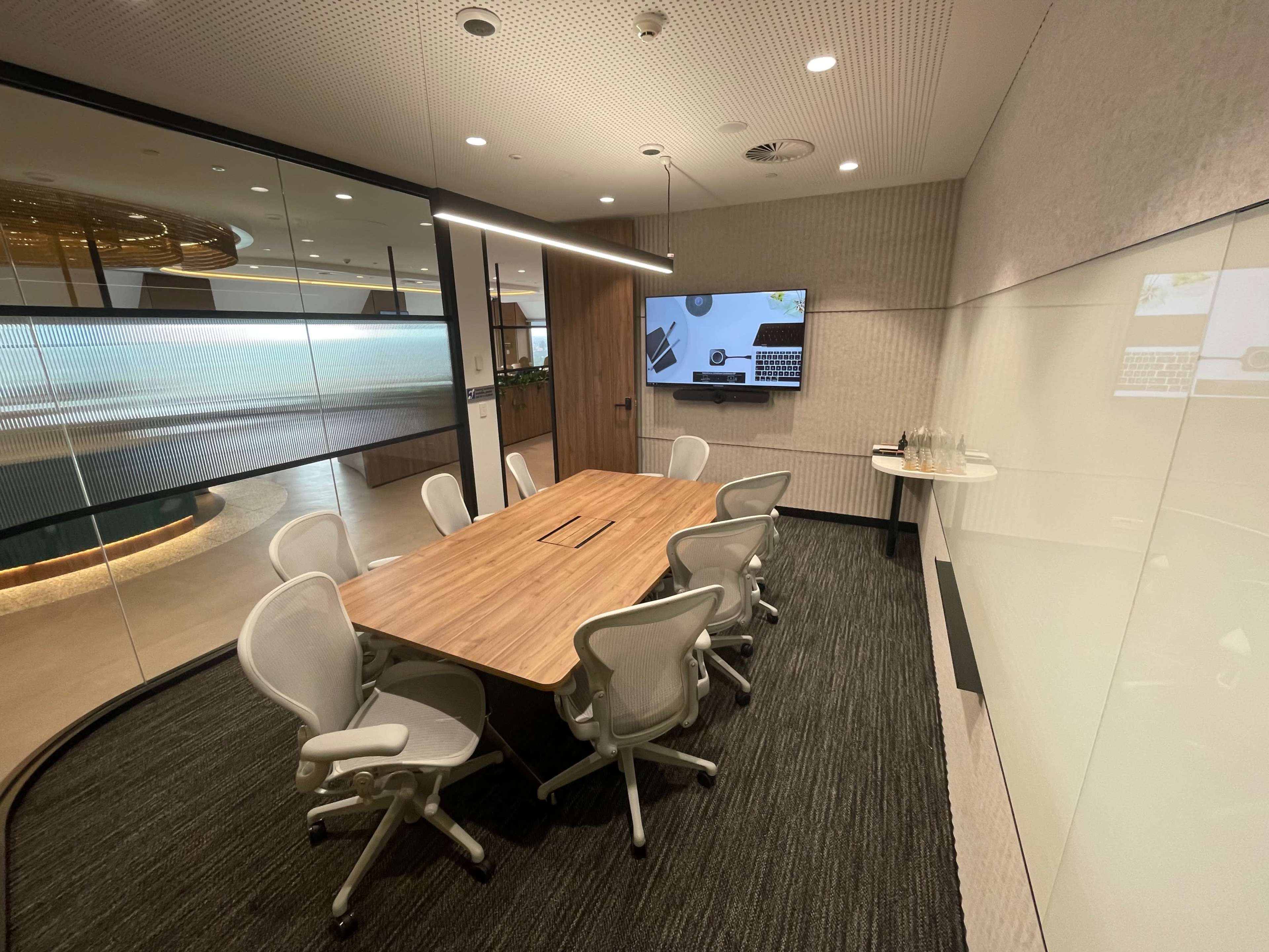 A modern conference room features a large wooden table surrounded by ergonomic chairs, with a wall-mounted screen displaying a digital interface.