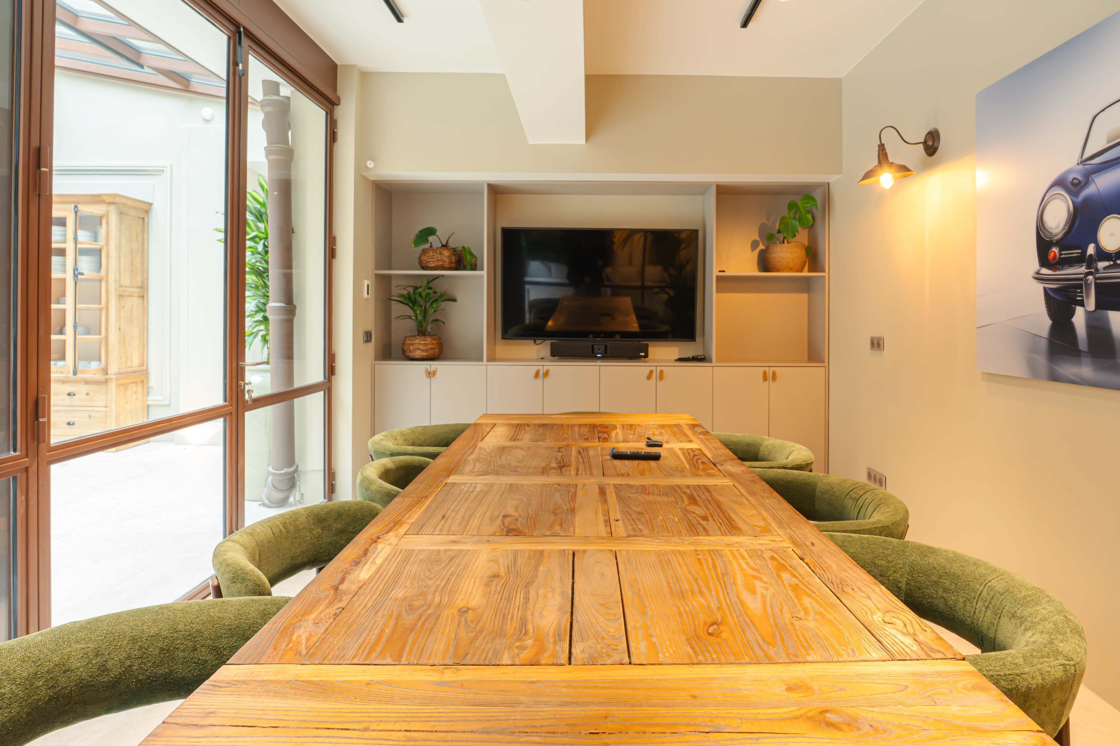 A long wooden dining table with green upholstered chairs is positioned in a well-lit room featuring a television and decorative shelves.