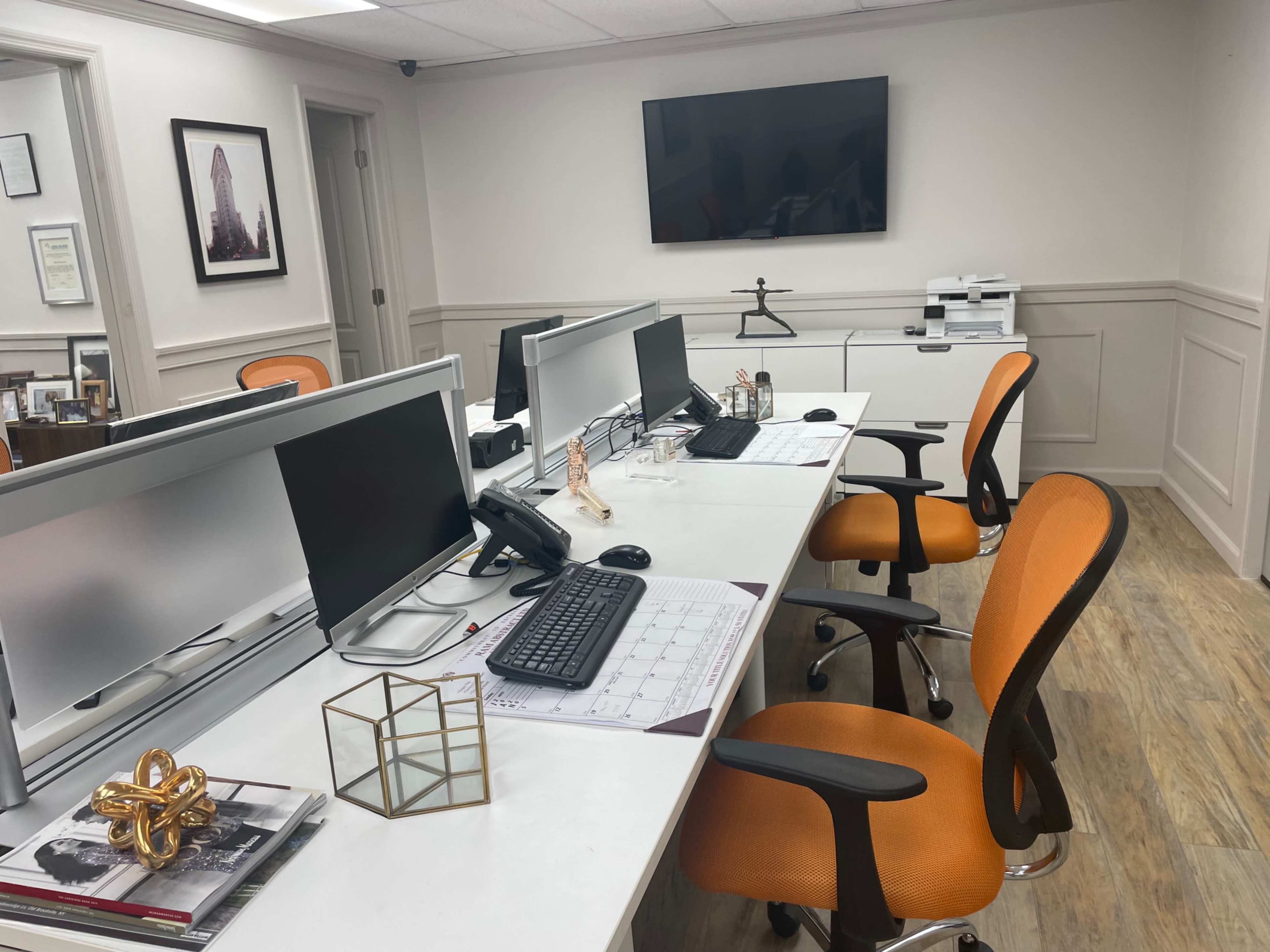 A modern office workspace with two computer desks, each equipped with monitors, keyboards, and orange chairs, arranged around a white table, with a television mounted on the wall and office supplies on the desks.