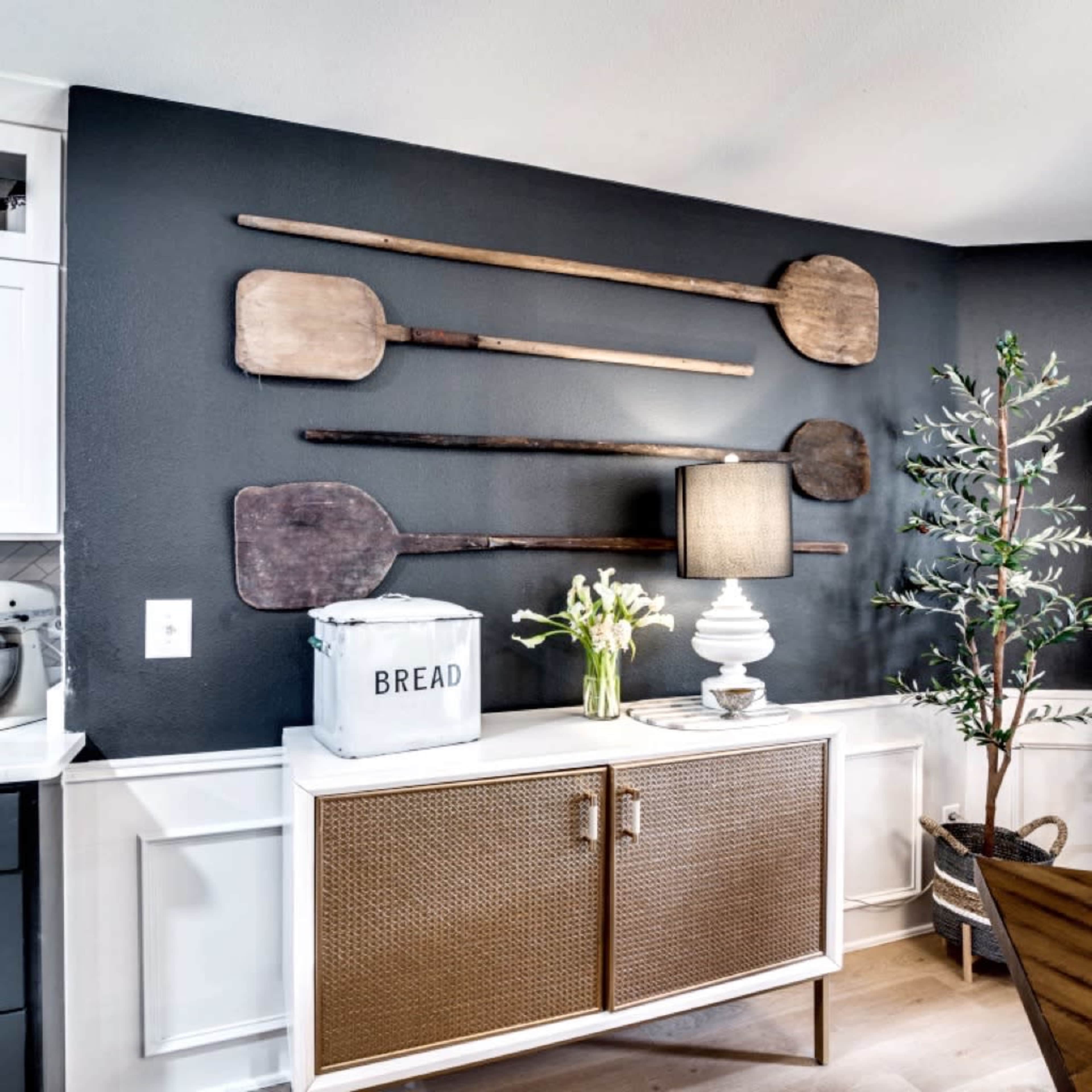 A collection of wooden paddles is mounted on a dark wall above a light cabinet, which holds a lamp and a decorative bread box.
