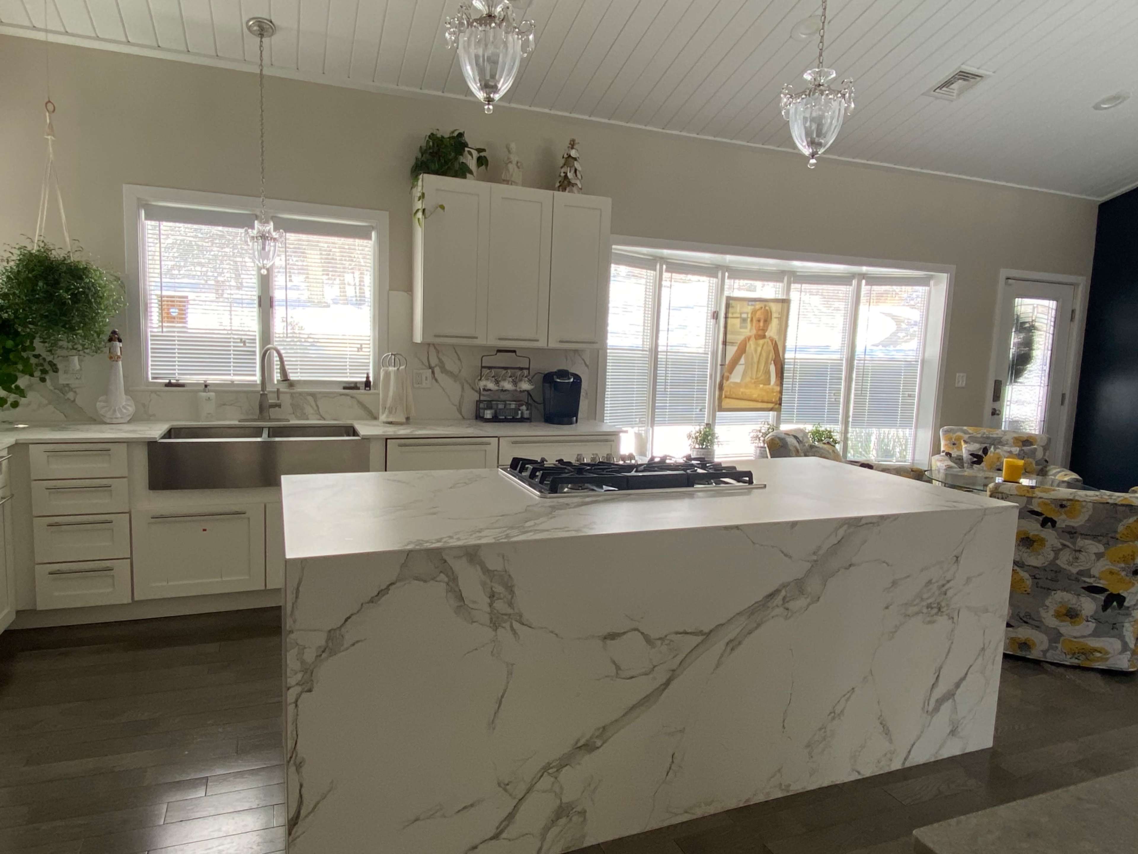A modern kitchen features a large marble island with a gas cooktop, white cabinetry, and ample natural light from large windows.