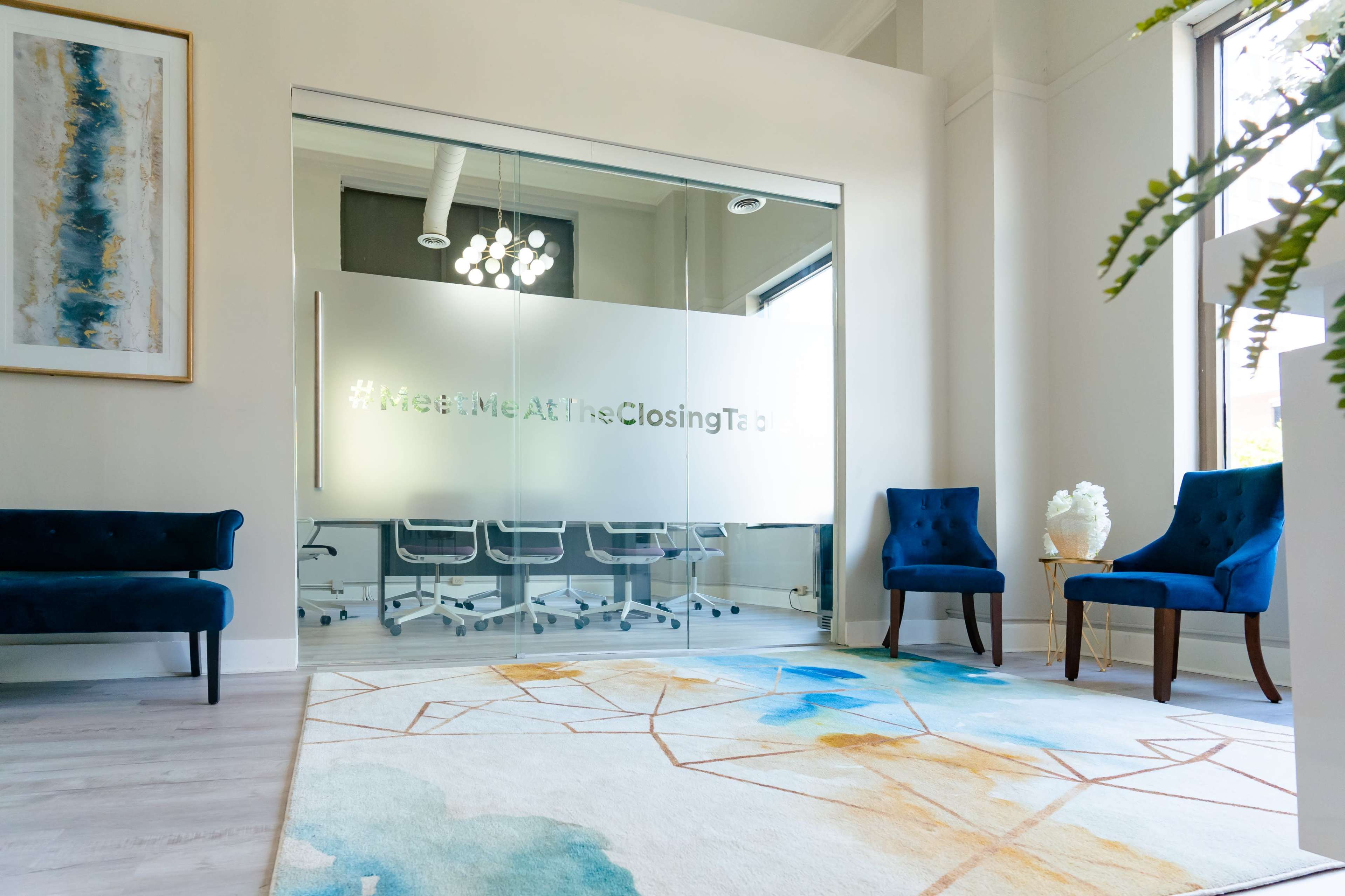 A modern office space with a glass meeting room, two blue chairs, and a colorful area rug on a light wood floor.