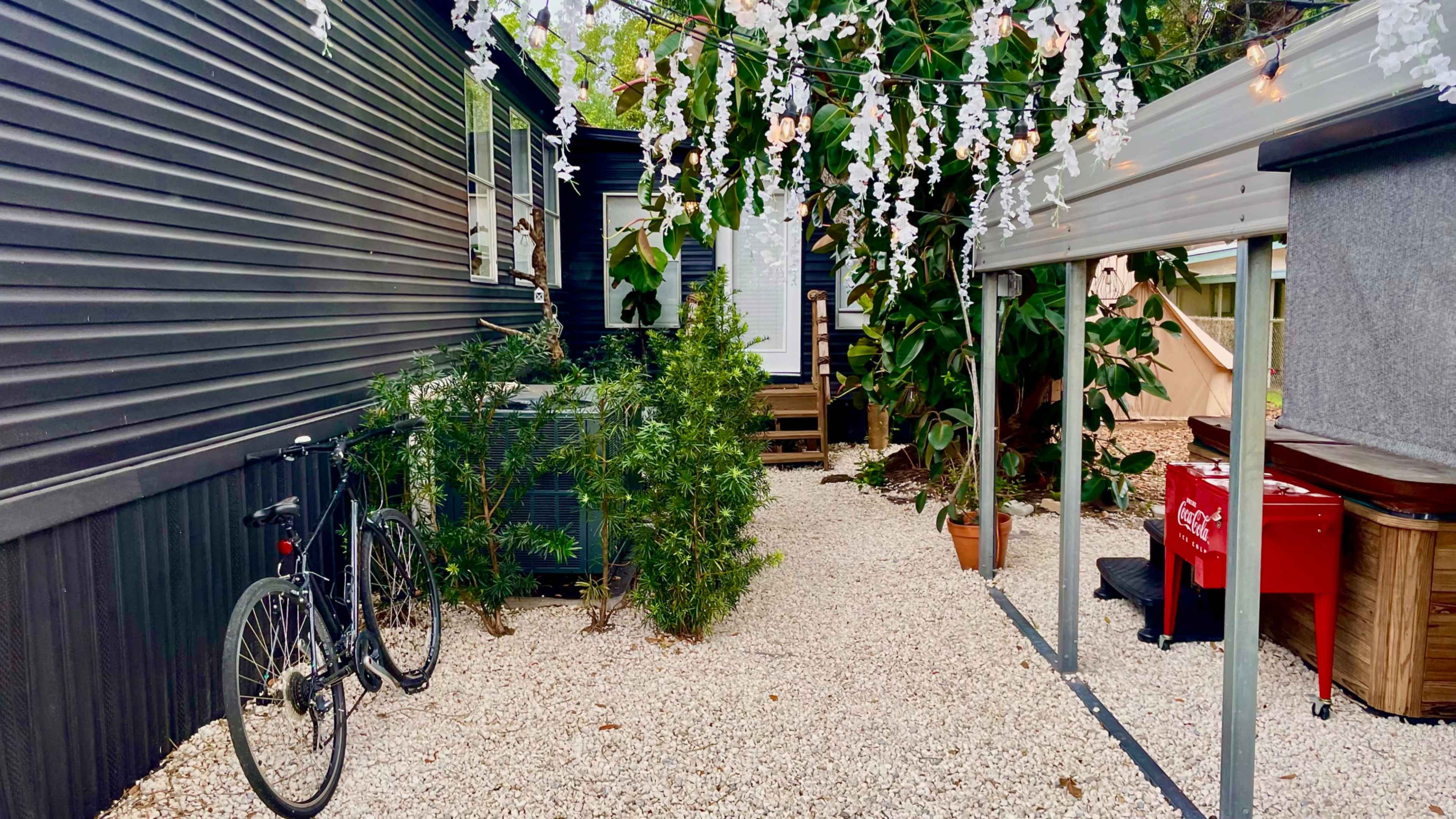 The scene shows a gravel pathway between a house and a canopy, with bicycles parked beside greenery and string lights overhead.