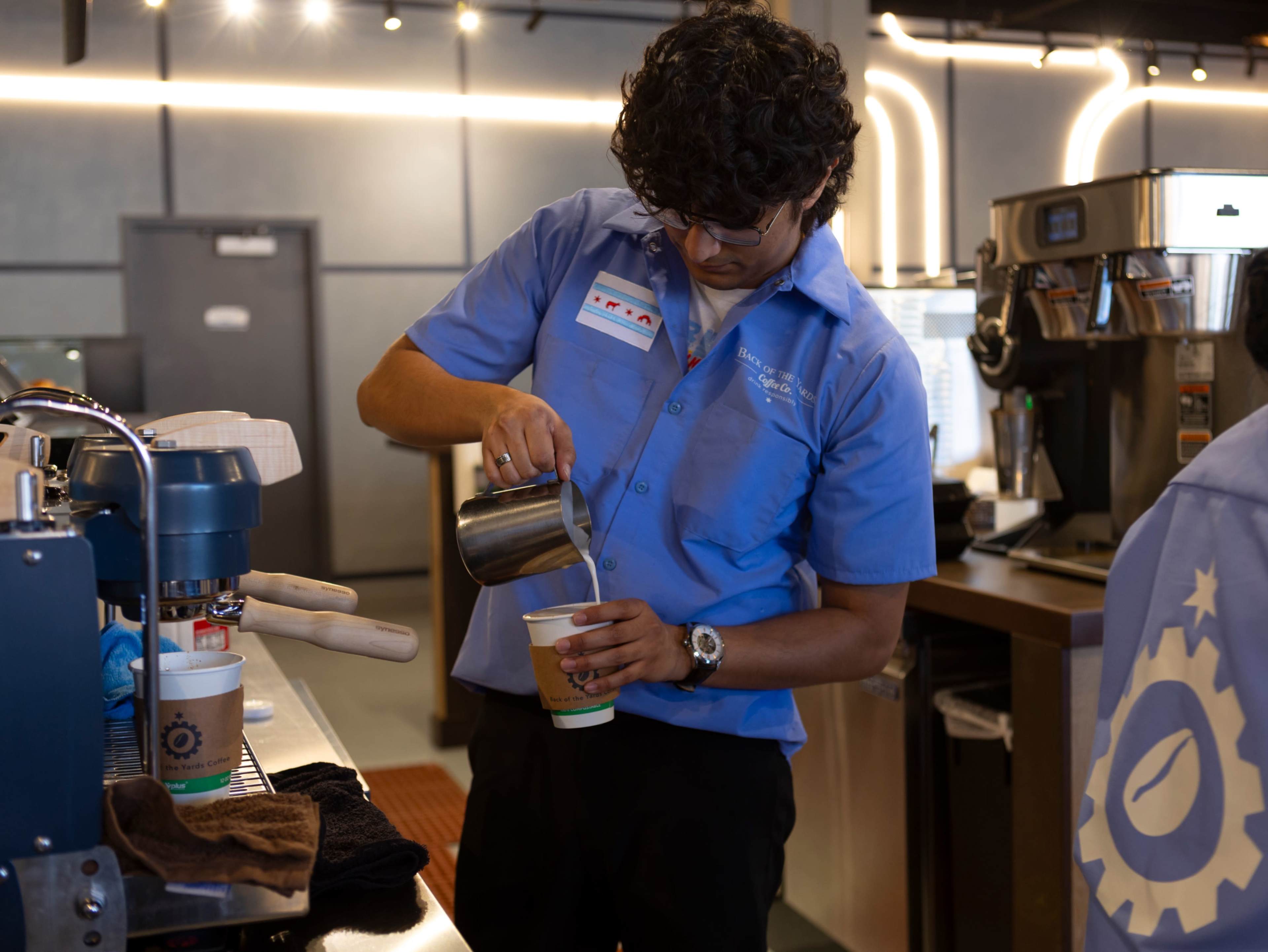A barista in a blue shirt pours steamed milk from a metal pitcher into a disposable coffee cup at a coffee shop.