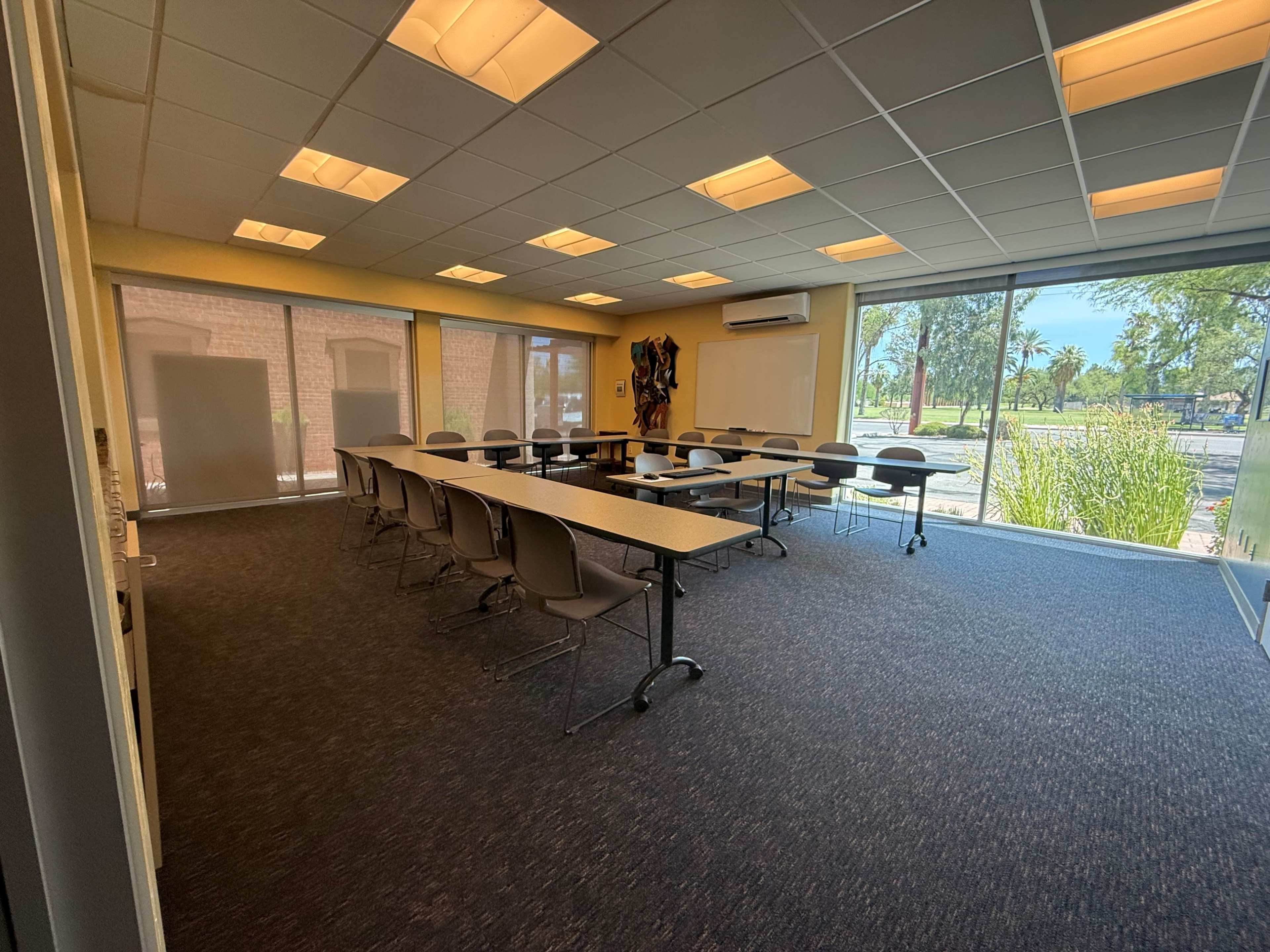 A spacious meeting room features a long table with chairs arranged around it, large windows allowing natural light, and a view of greenery outside.
