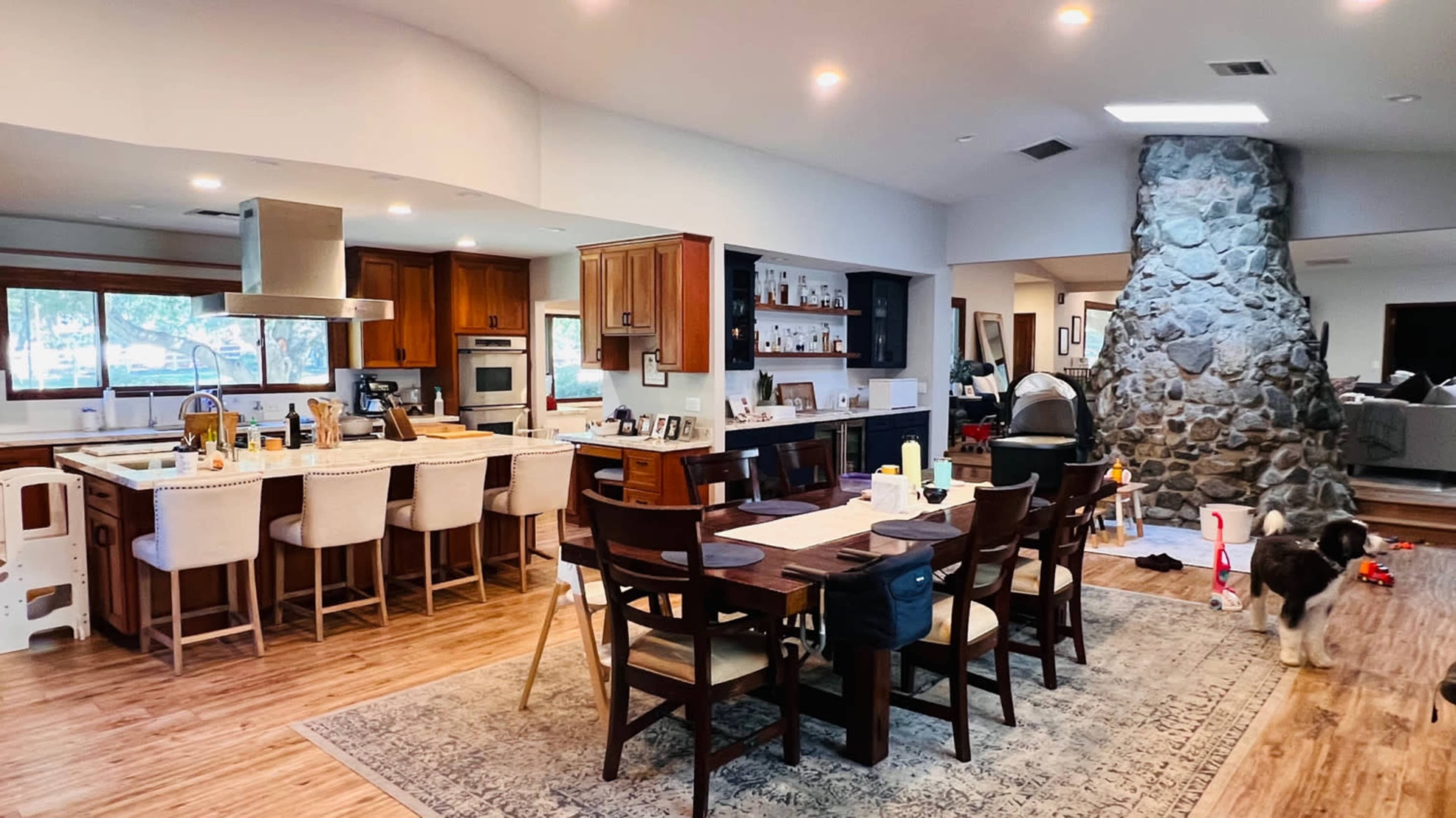 A spacious kitchen and dining area featuring wooden cabinets, a large stone fireplace, a dining table, and a dog on a rug.