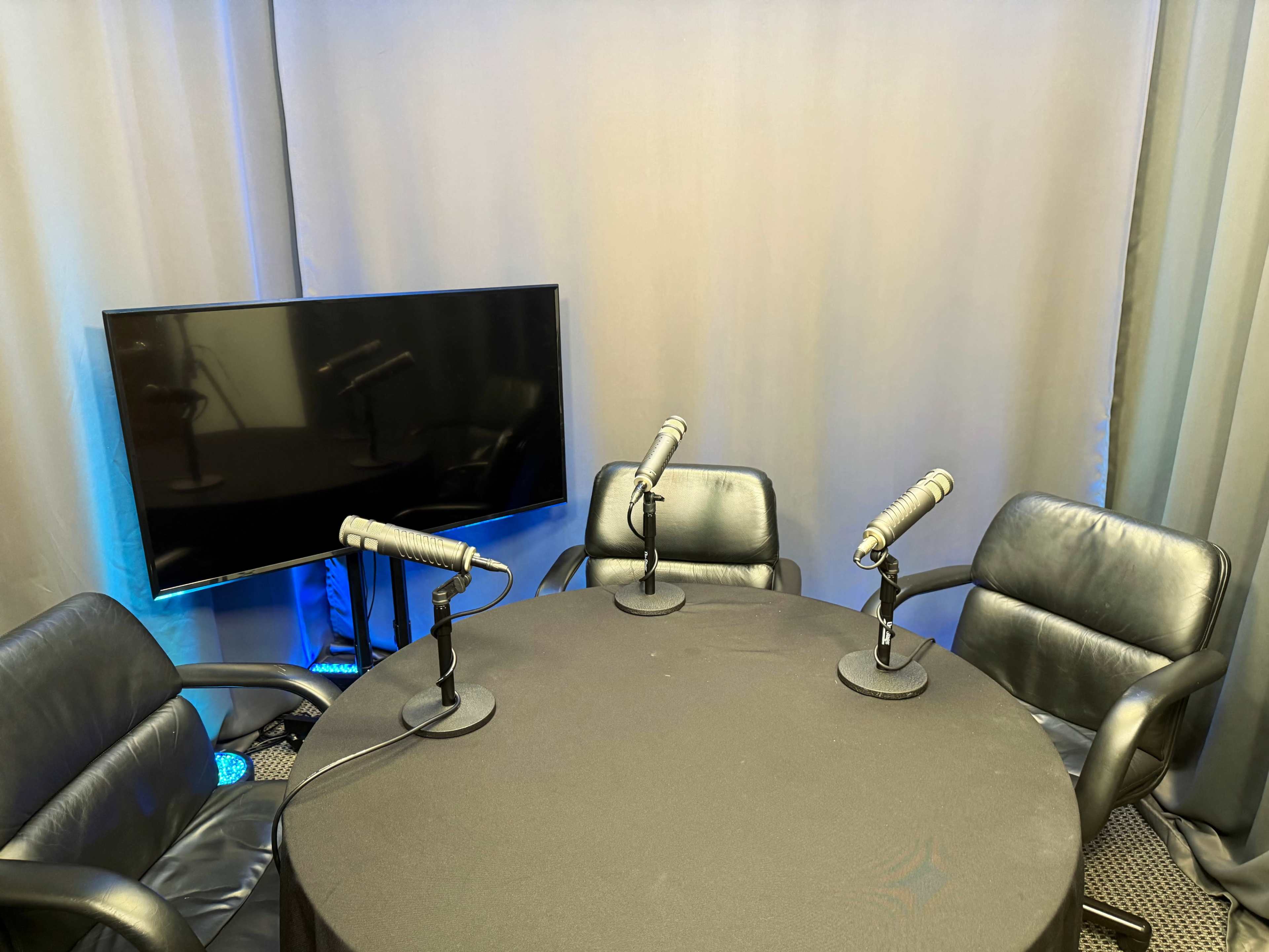 The image shows a small podcast setup featuring a round table with three microphones, three black leather chairs, and a television screen against a gray curtain backdrop.