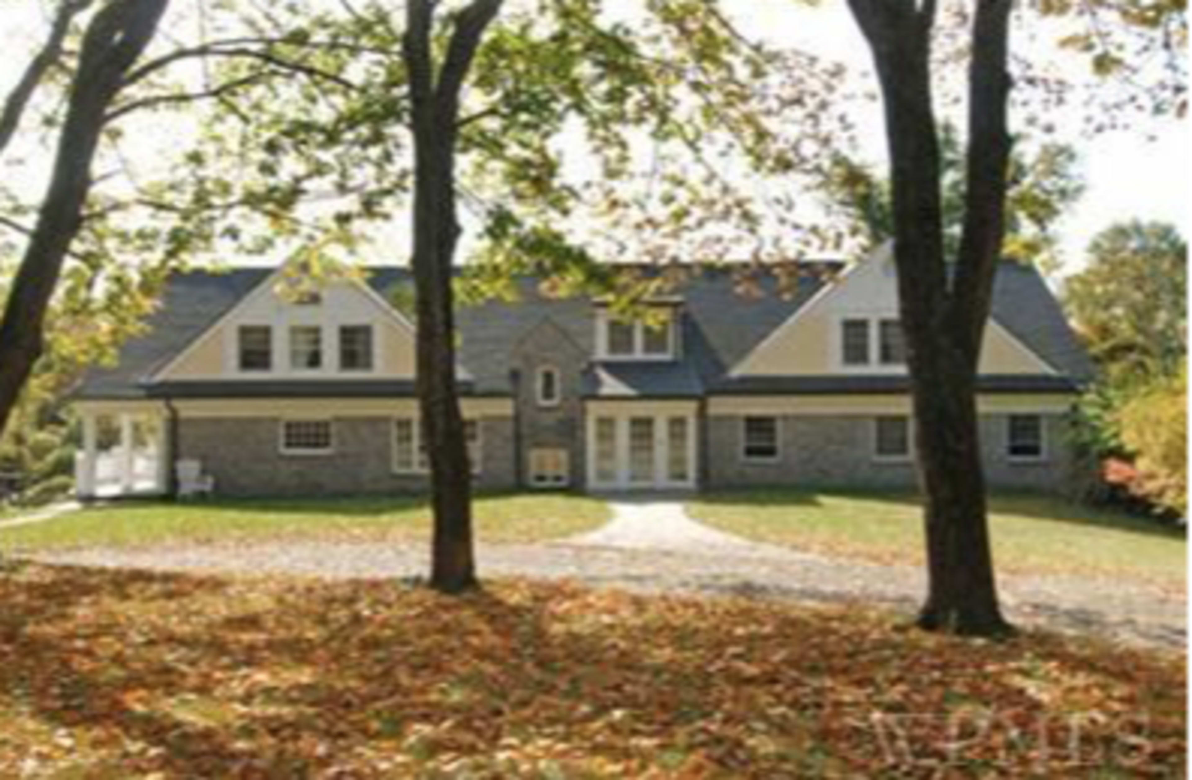 A large stone house with a gray roof sits in a yard covered in fallen leaves, surrounded by trees.