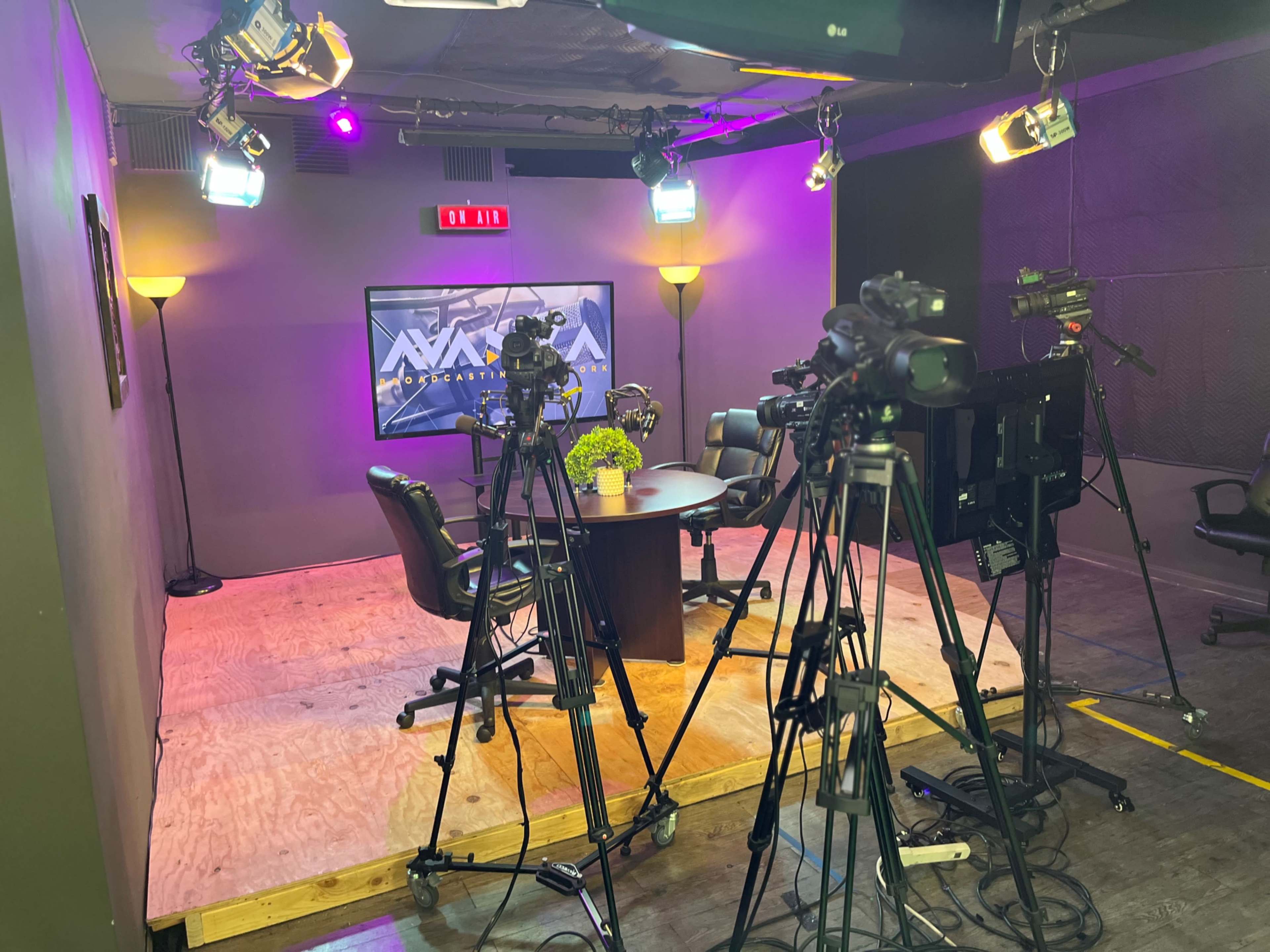 A television studio setup with multiple cameras positioned around a round table surrounded by four chairs and illuminated by various lights.