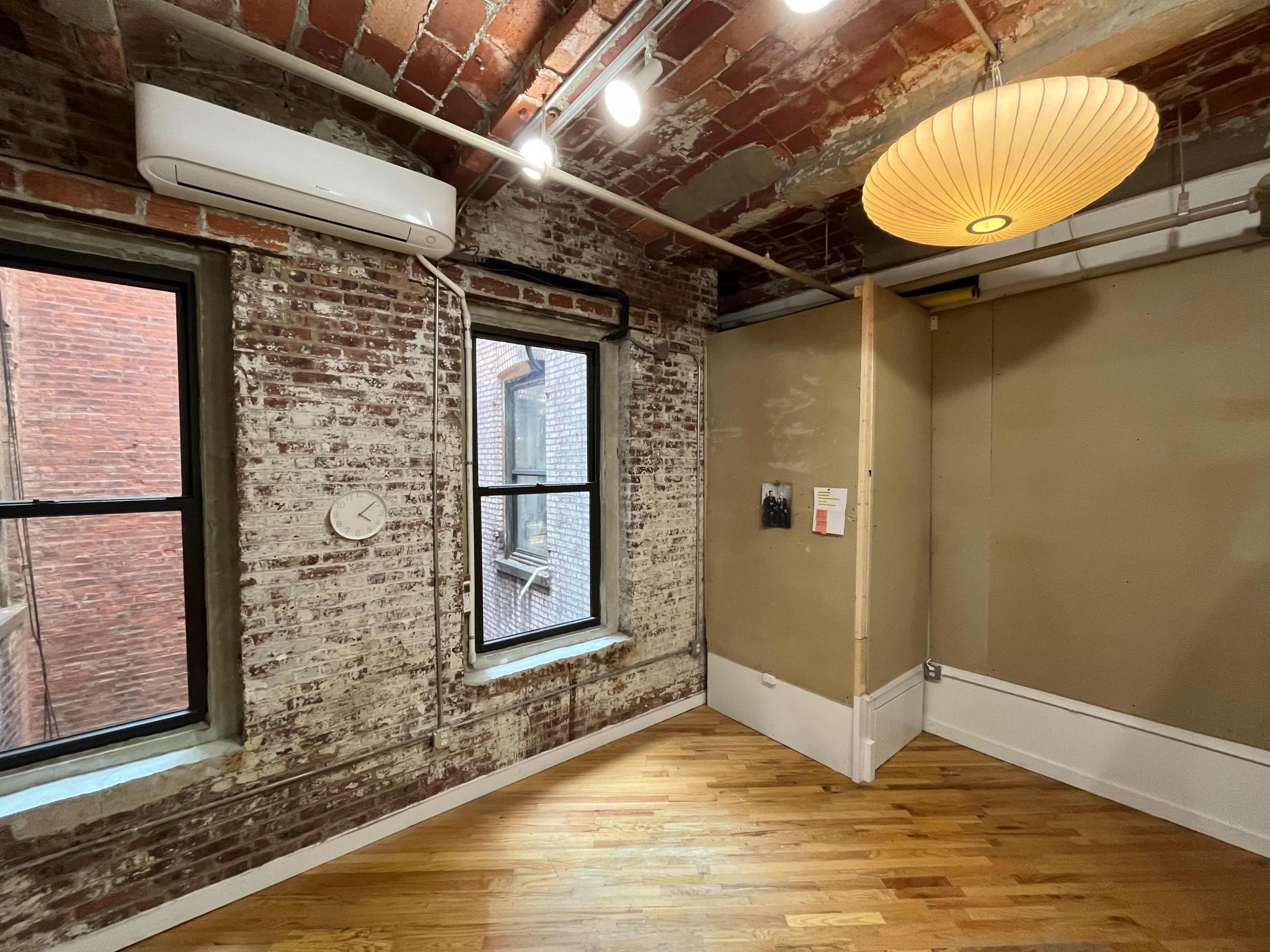 A room with exposed brick walls, two windows, wooden flooring, and a round pendant light.
