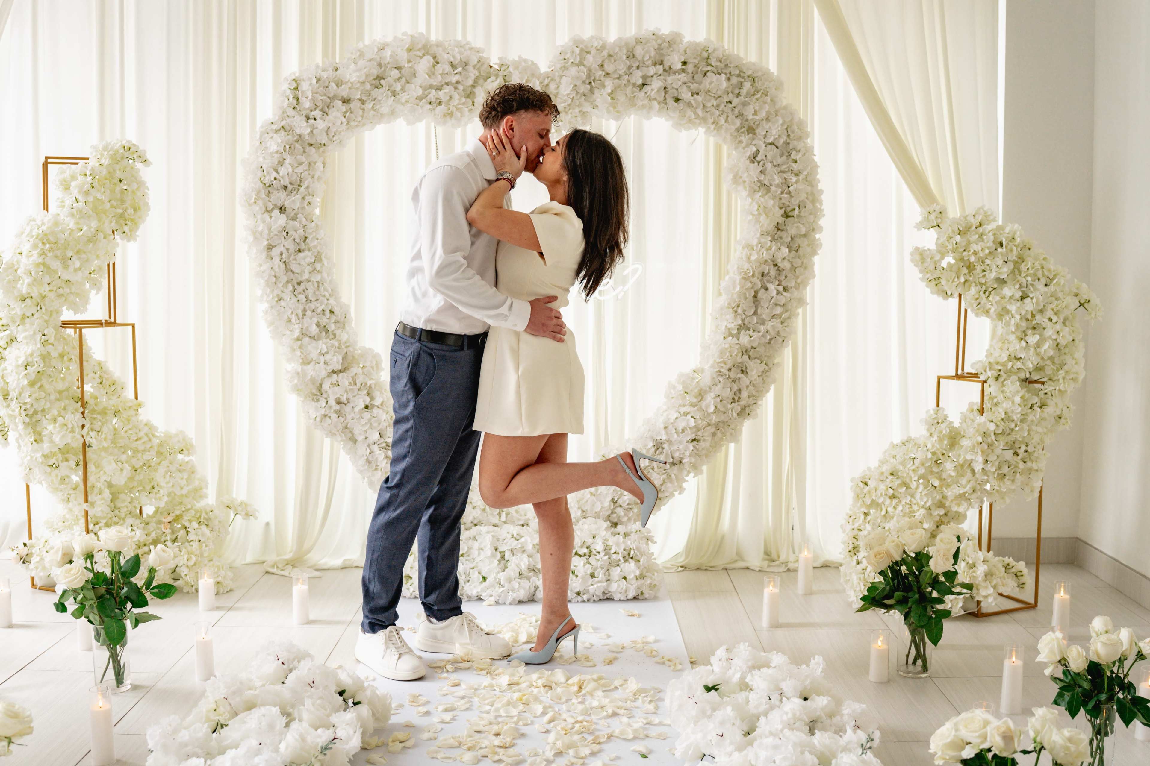 A couple embraces in front of a heart-shaped floral arrangement with white flowers, surrounded by candles and rose petals on the floor.