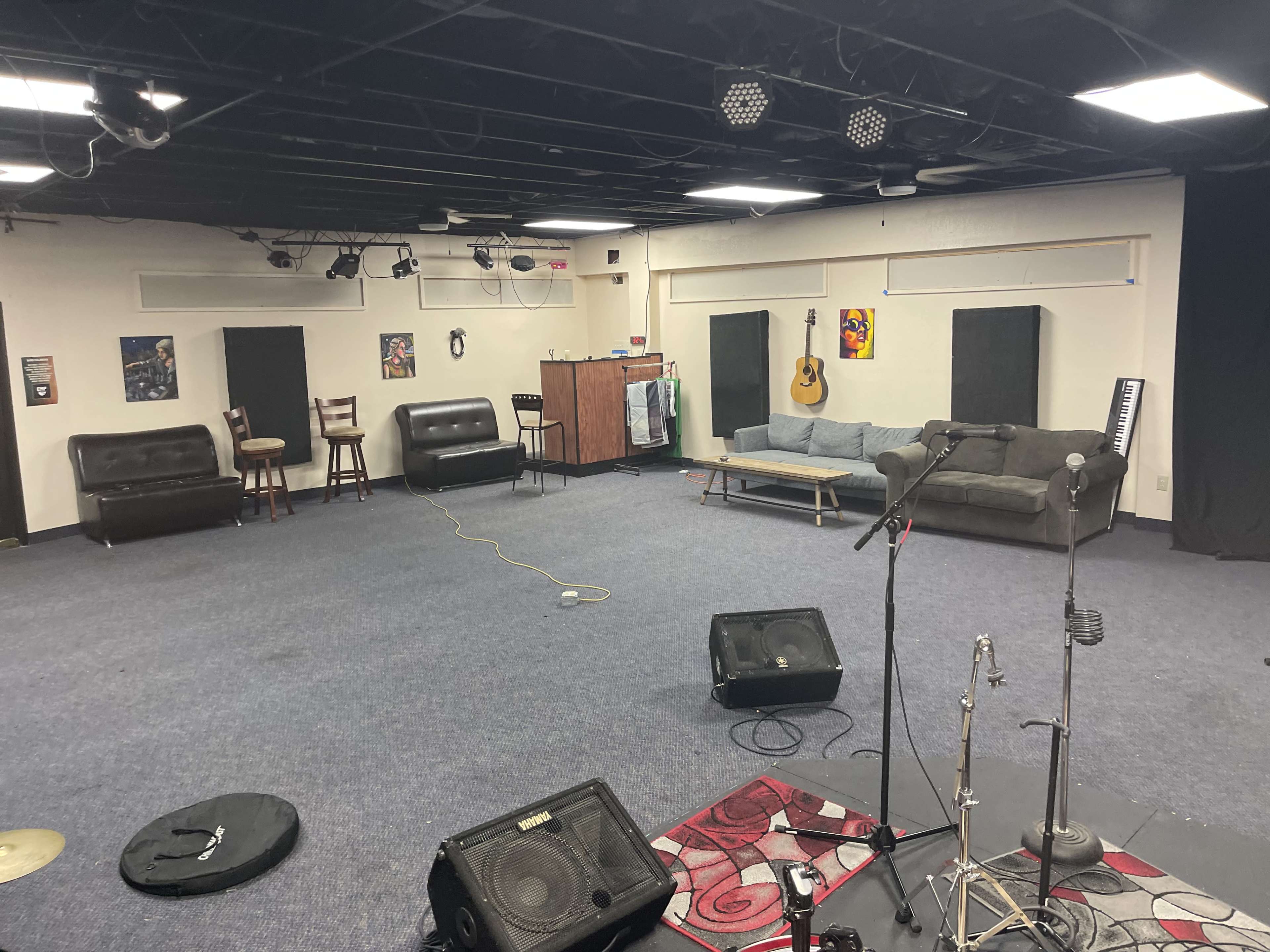 The image shows a spacious rehearsal studio with black walls, a few instruments on the floor, sofas along the sides, and a small kitchenette area in the background.