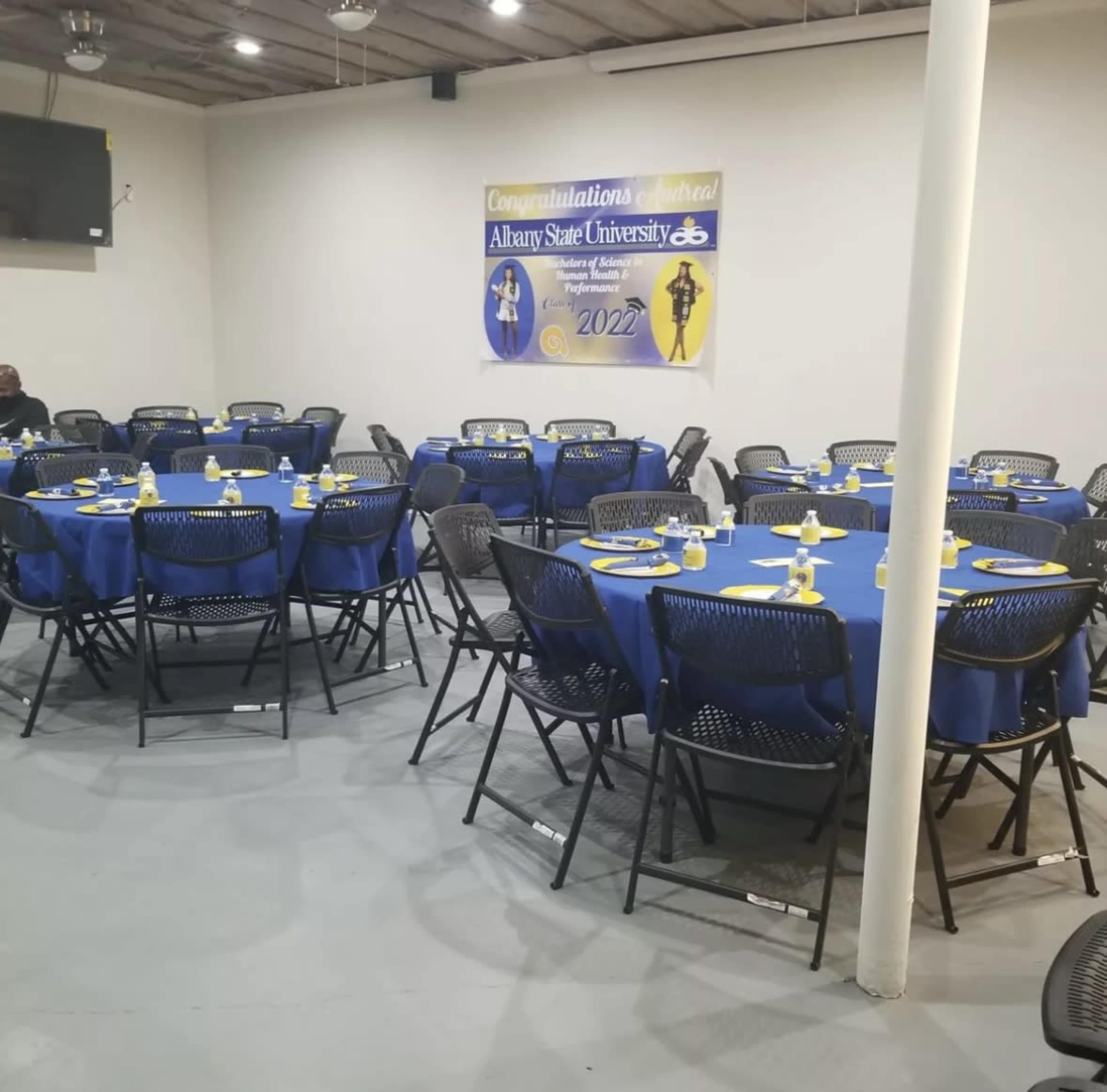 A banquet hall is set up with round tables covered in blue tablecloths, each with yellow napkins, and a banner celebrating Albany State University in the background.