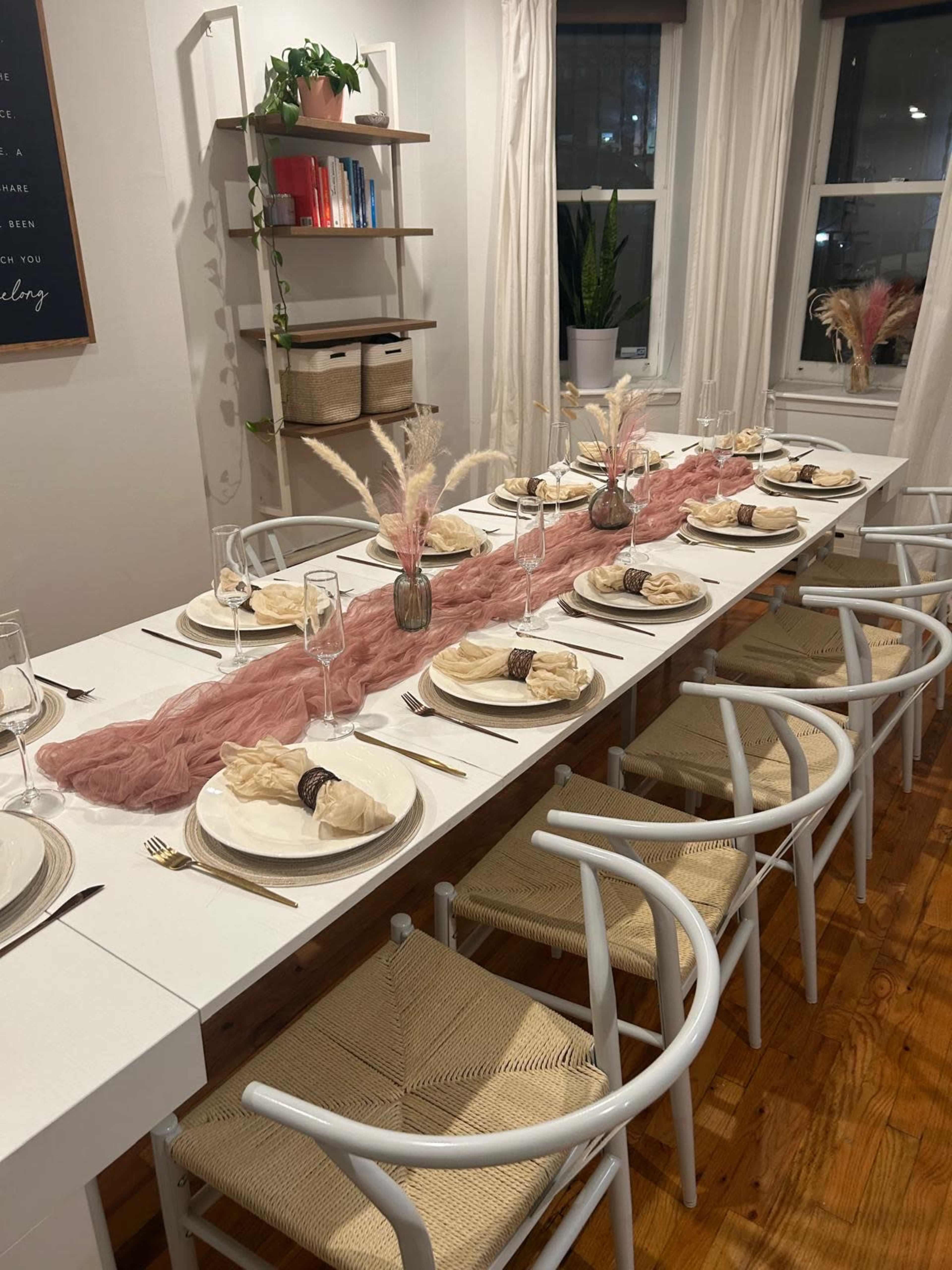 A long dining table is set with plates, glasses, and decorative elements, featuring a pink table runner and surrounded by woven chairs.