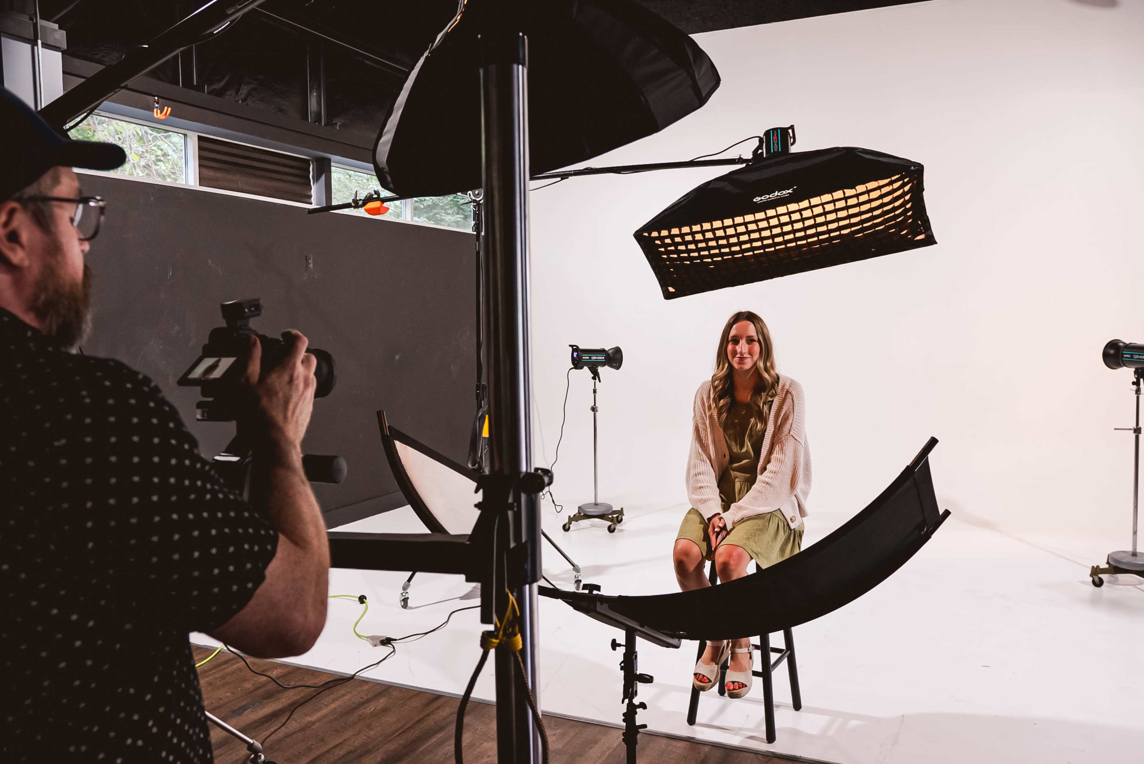 A photographer takes a portrait of a woman seated on a stool in a studio with a plain backdrop and multiple lighting devices.