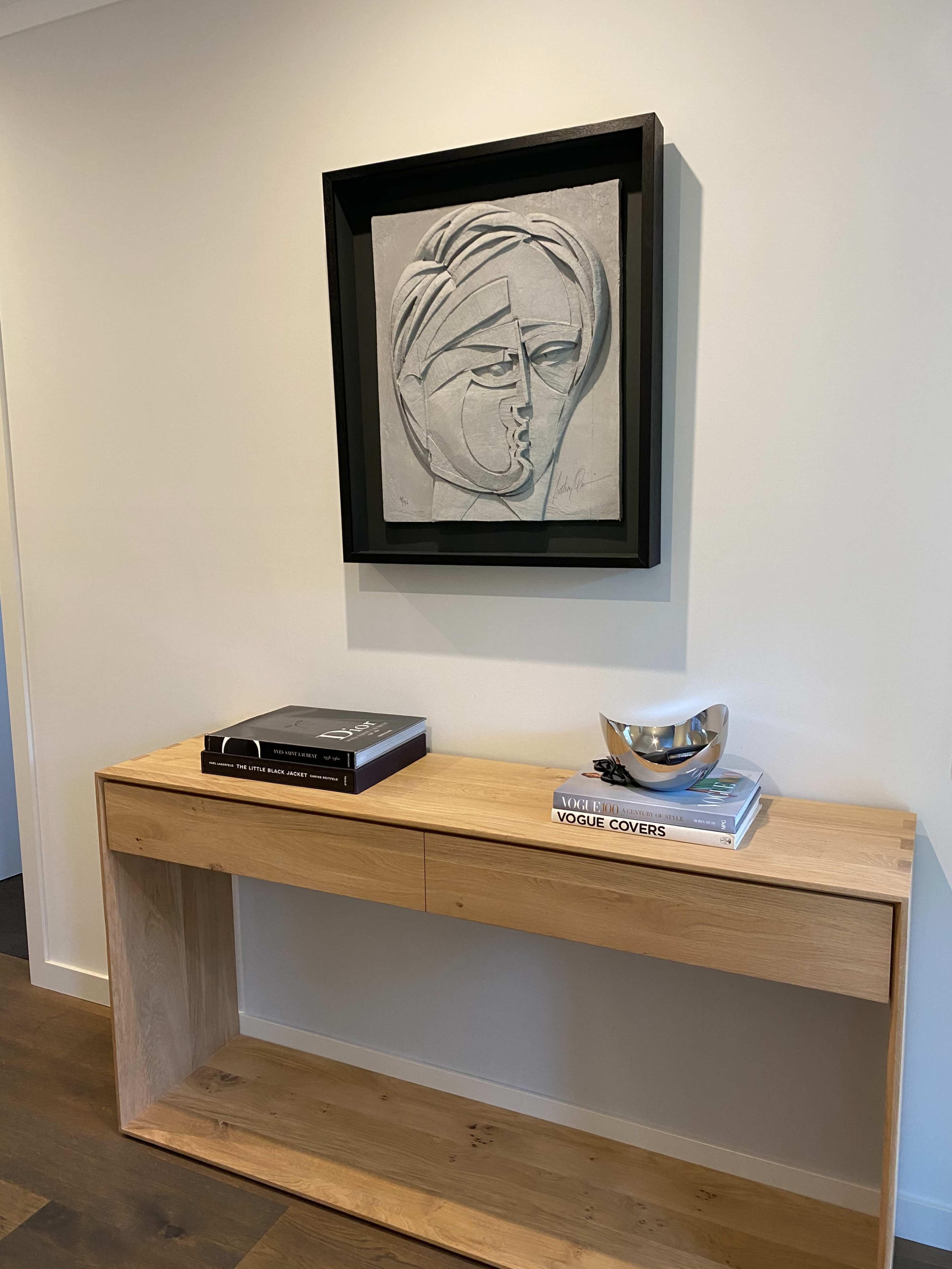 A light-colored wall features a framed abstract artwork above a wooden console table, which holds a stack of books and a decorative bowl.