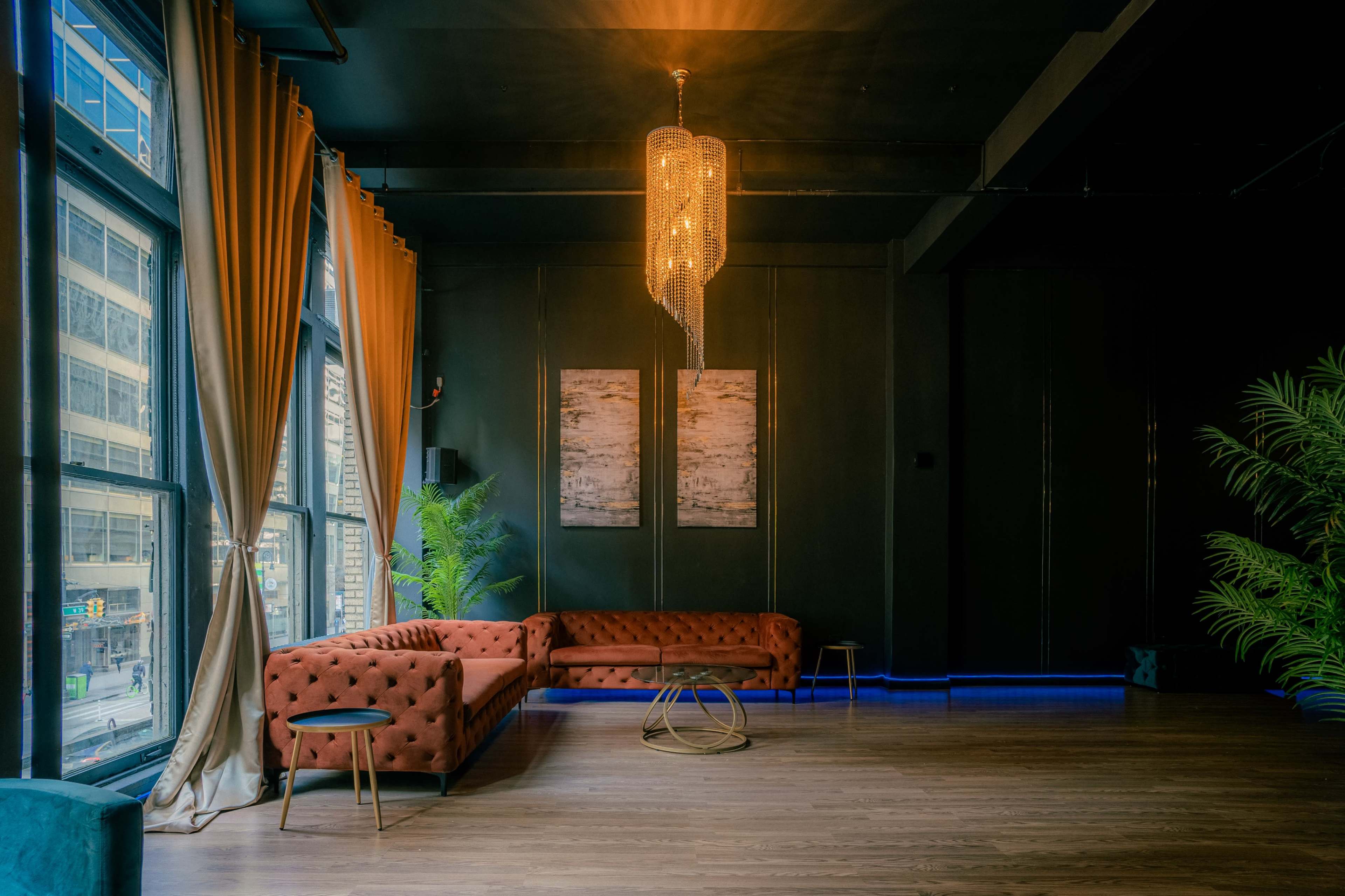A stylish lounge area with a large orange couch, a round table, and a chandelier, illuminated by natural light from large windows.