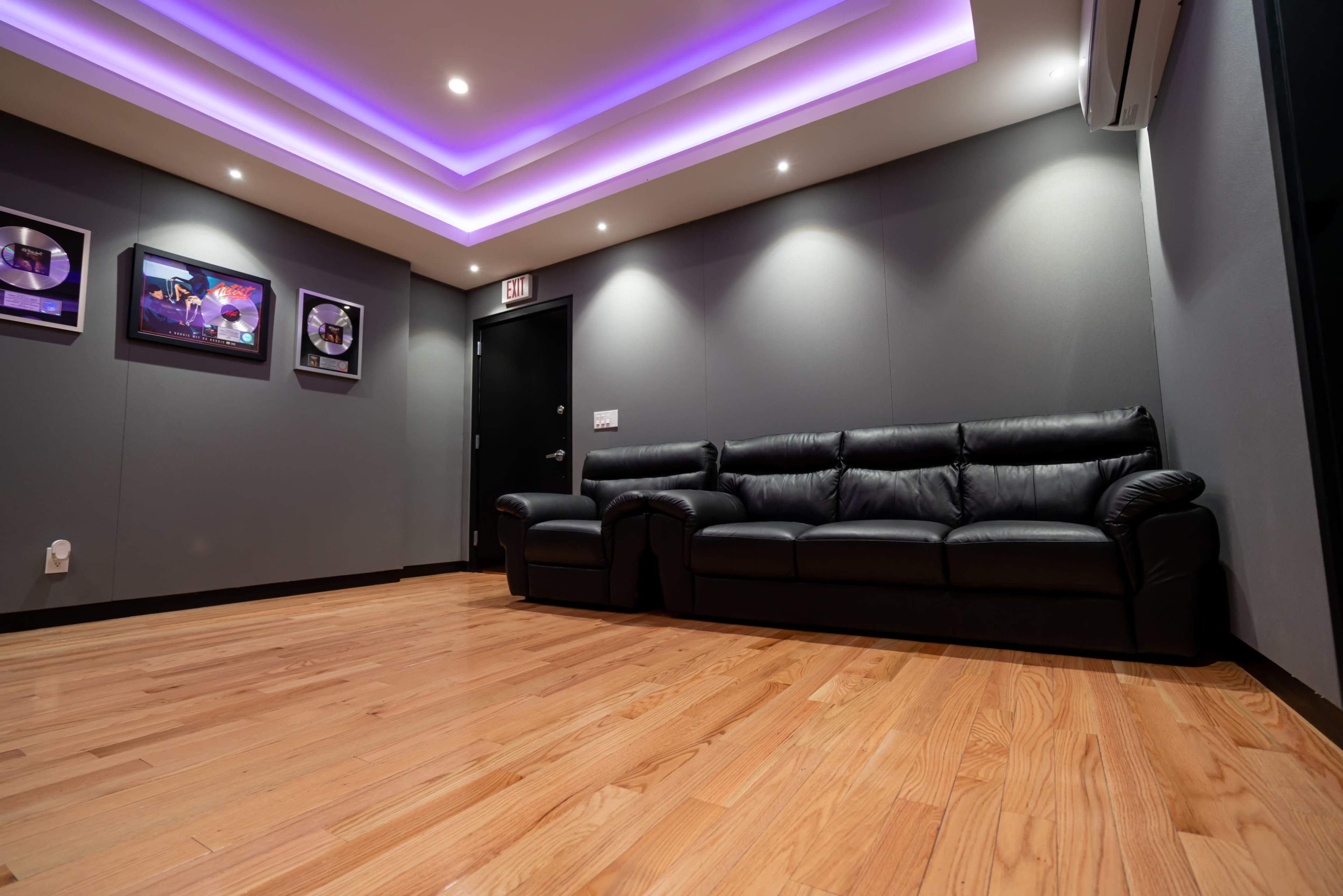 A room with hardwood flooring, featuring a black leather couch against a gray wall with framed music awards and purple lighting in the ceiling.