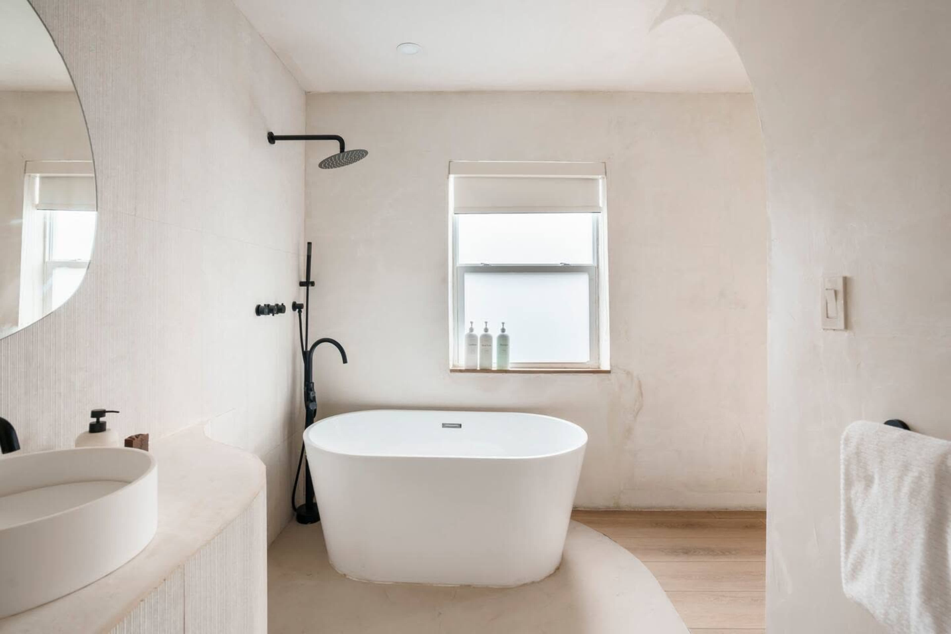The bathroom features a freestanding white bathtub, a round mirror, and a minimalist design with neutral colored walls and wooden flooring.