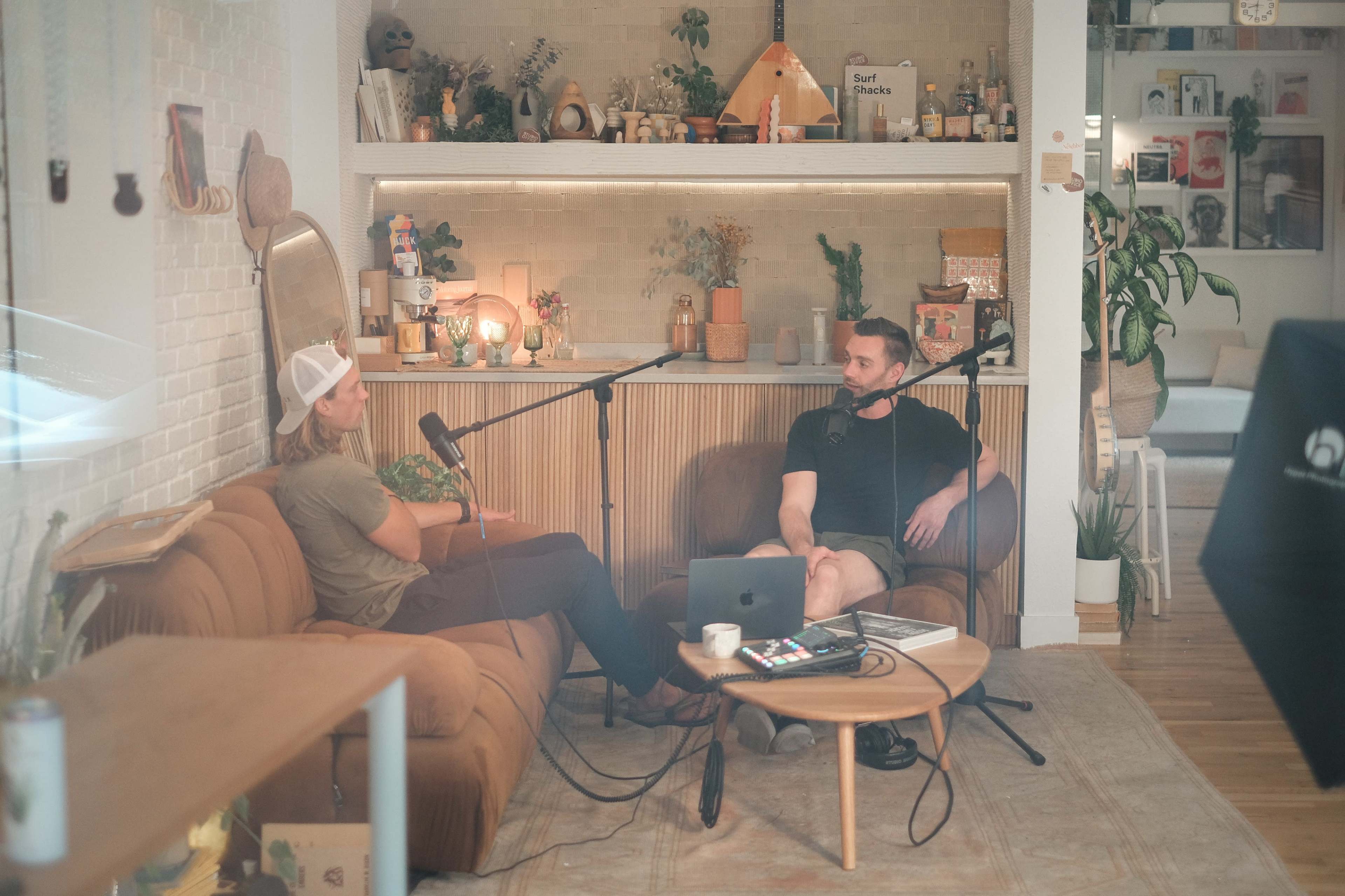 Two people are seated on a couch in a cozy studio, discussing into microphones while a laptop and various decorative items are visible around them.