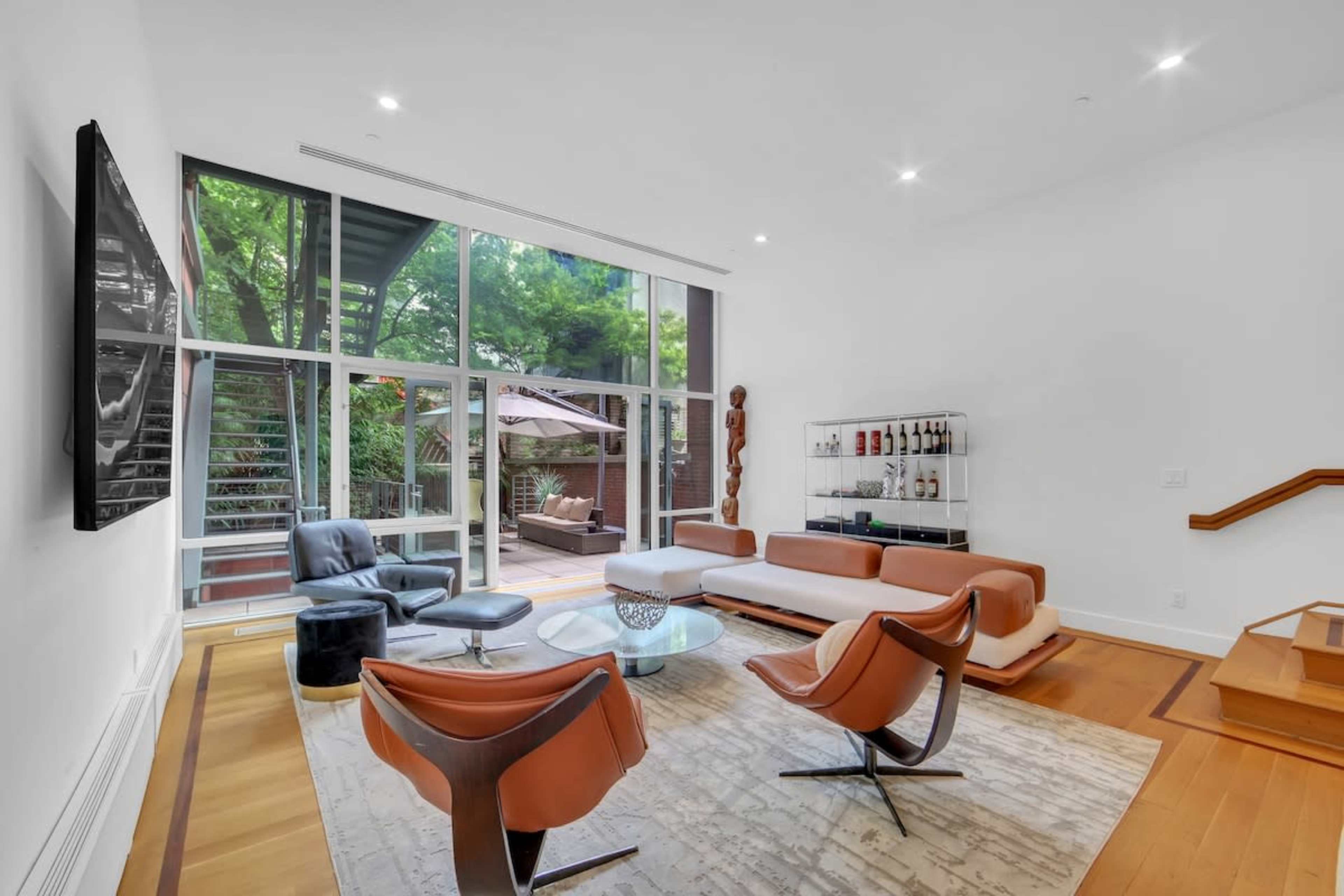 A modern living room features large windows overlooking a patio, minimalist furniture, and hardwood flooring.