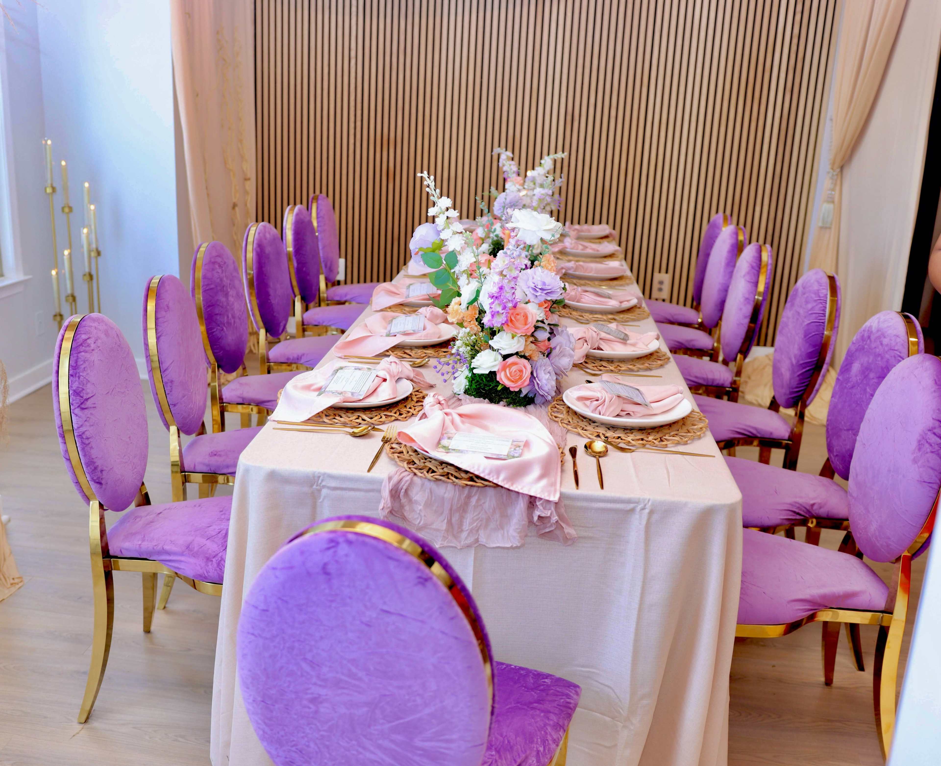 A long dining table is set with pink and gold chairs, adorned with a floral centerpiece and elegant tableware.