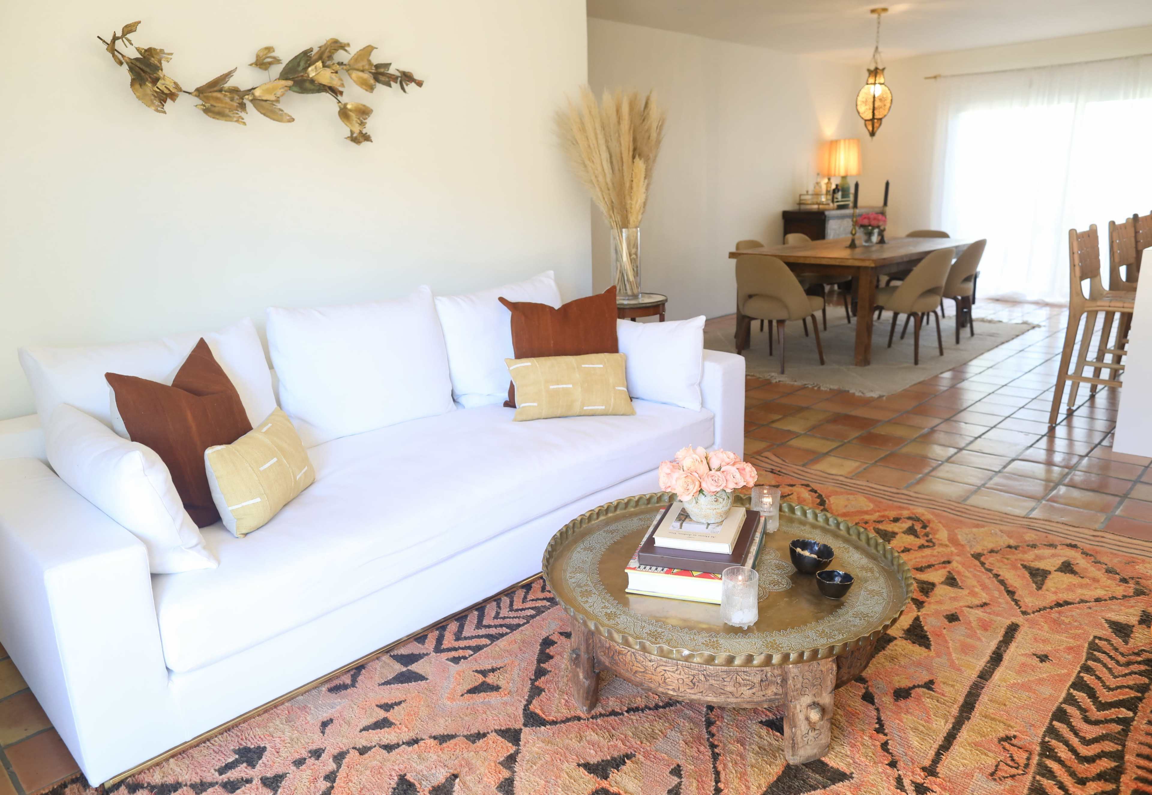 A living room with a white sofa adorned with decorative pillows, a patterned rug, a wooden coffee table, and a dining area in the background featuring a round table and chairs.