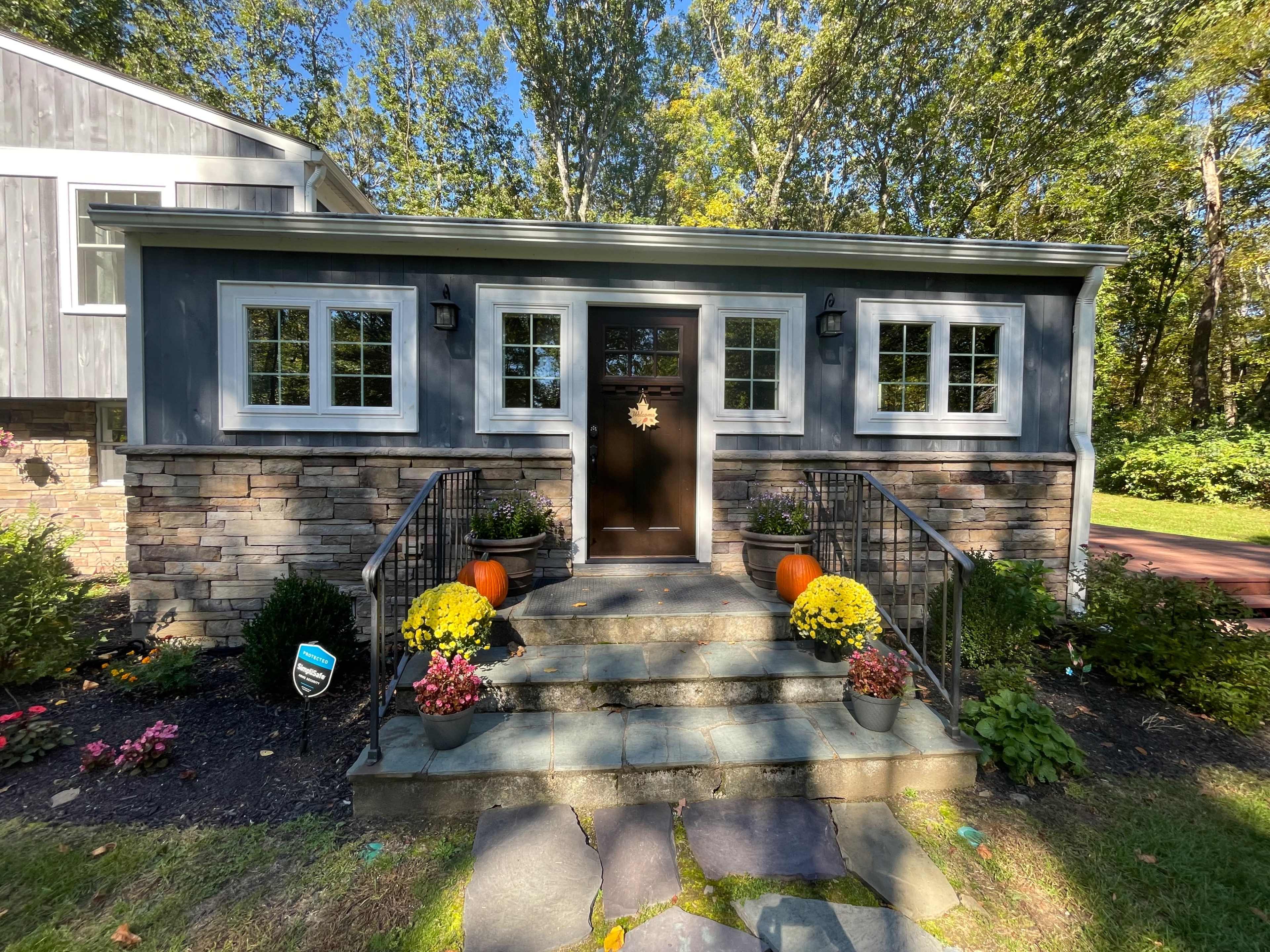 The image shows a home entrance with a dark front door, flanked by large windows, surrounded by stone steps and colorful potted plants.