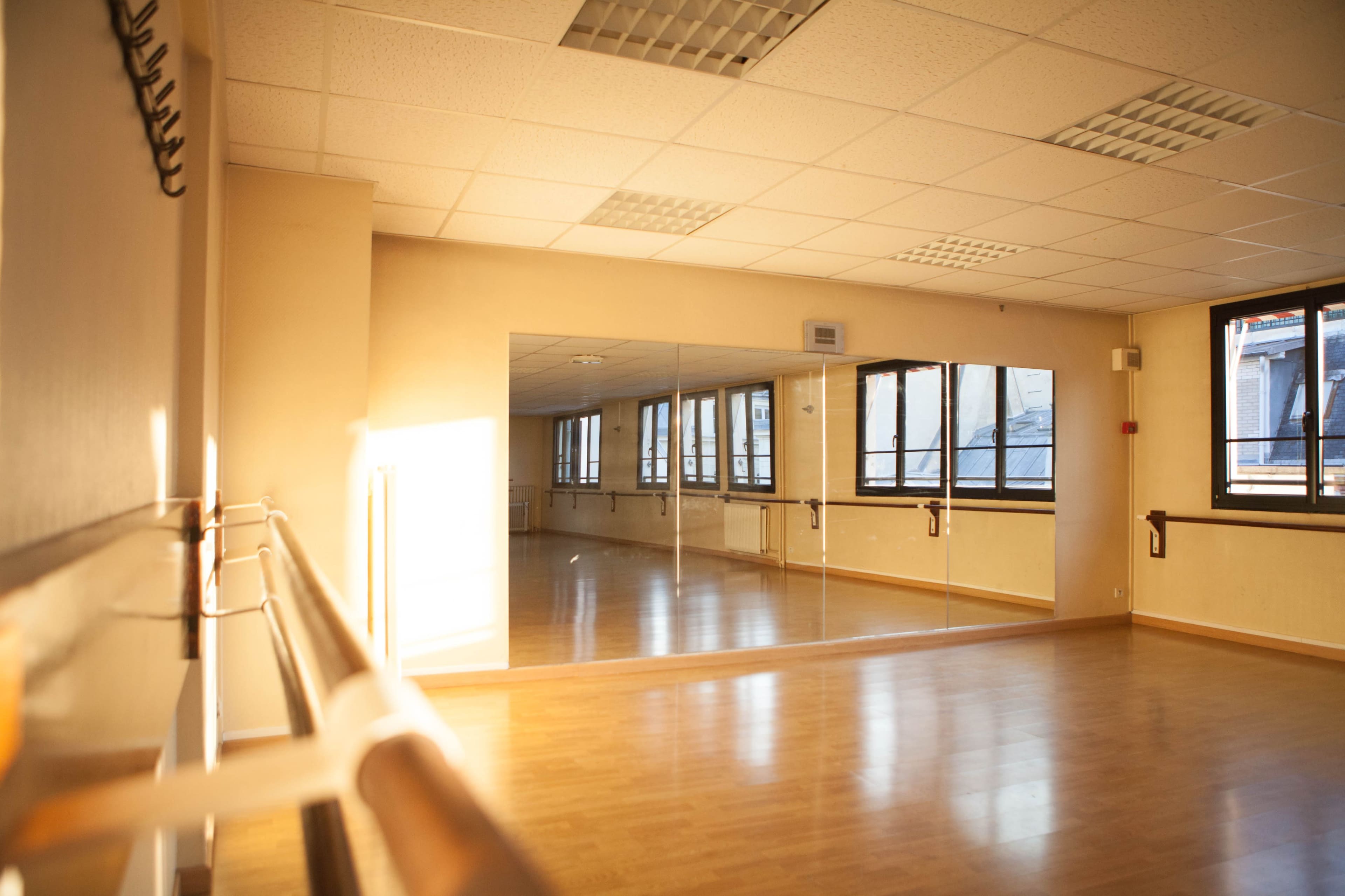 The image shows a bright dance studio with wooden flooring, large windows, and mirrored walls.