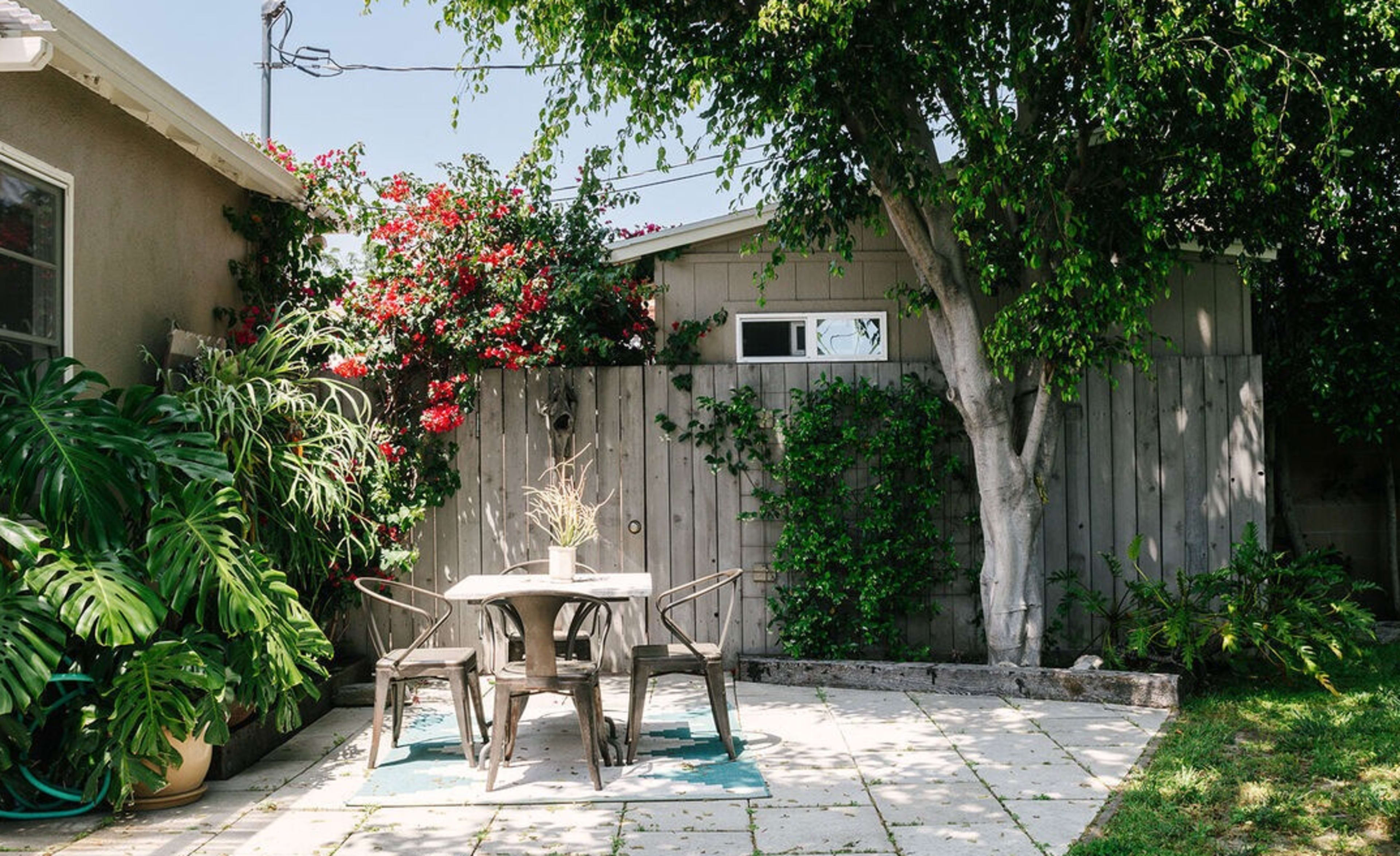 A small outdoor patio with a table and chairs is surrounded by greenery and flowering plants against a wooden fence.