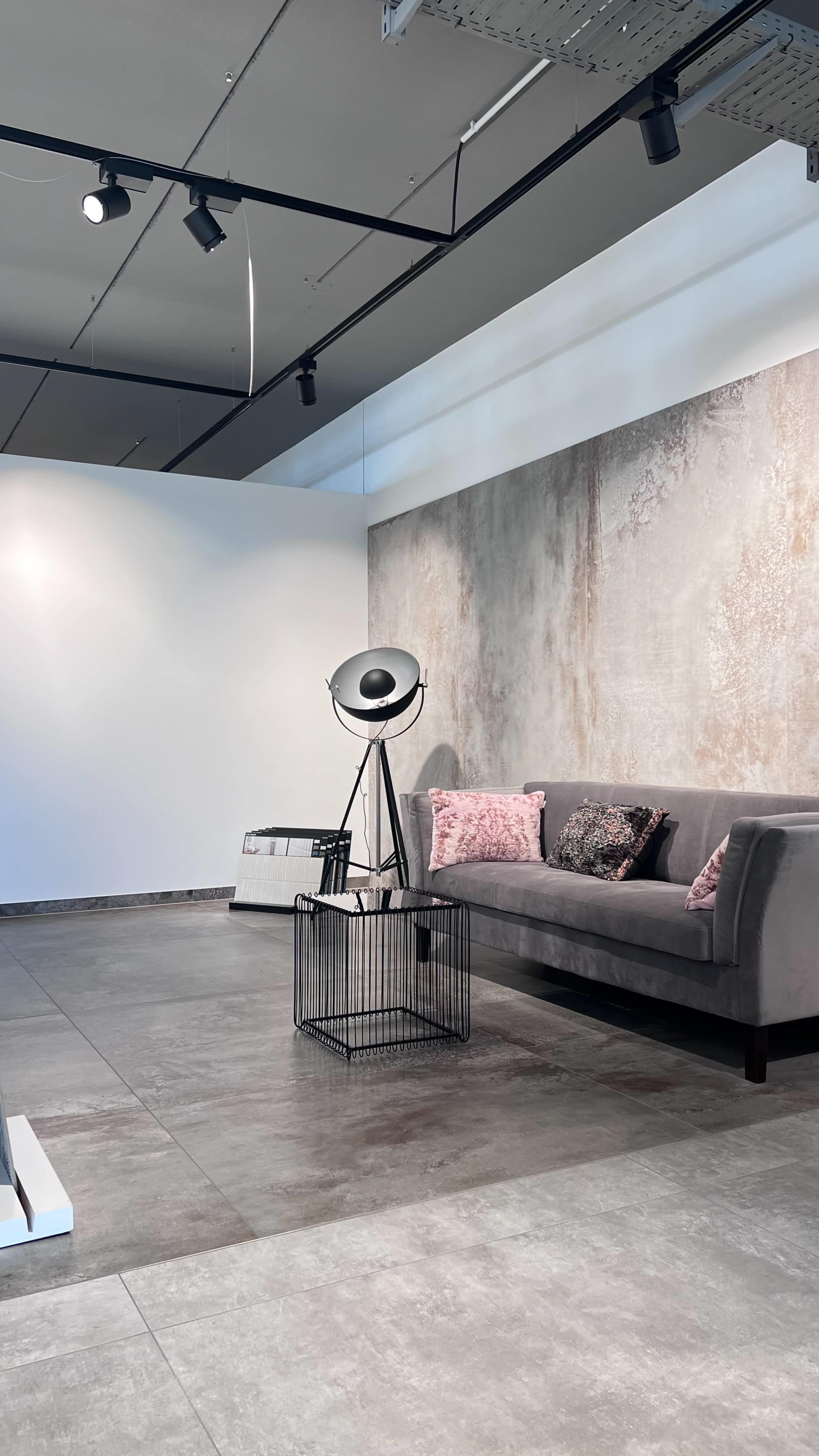 A gray sofa with decorative pillows is positioned next to a decorative lamp on a wire table in a minimalist interior space.