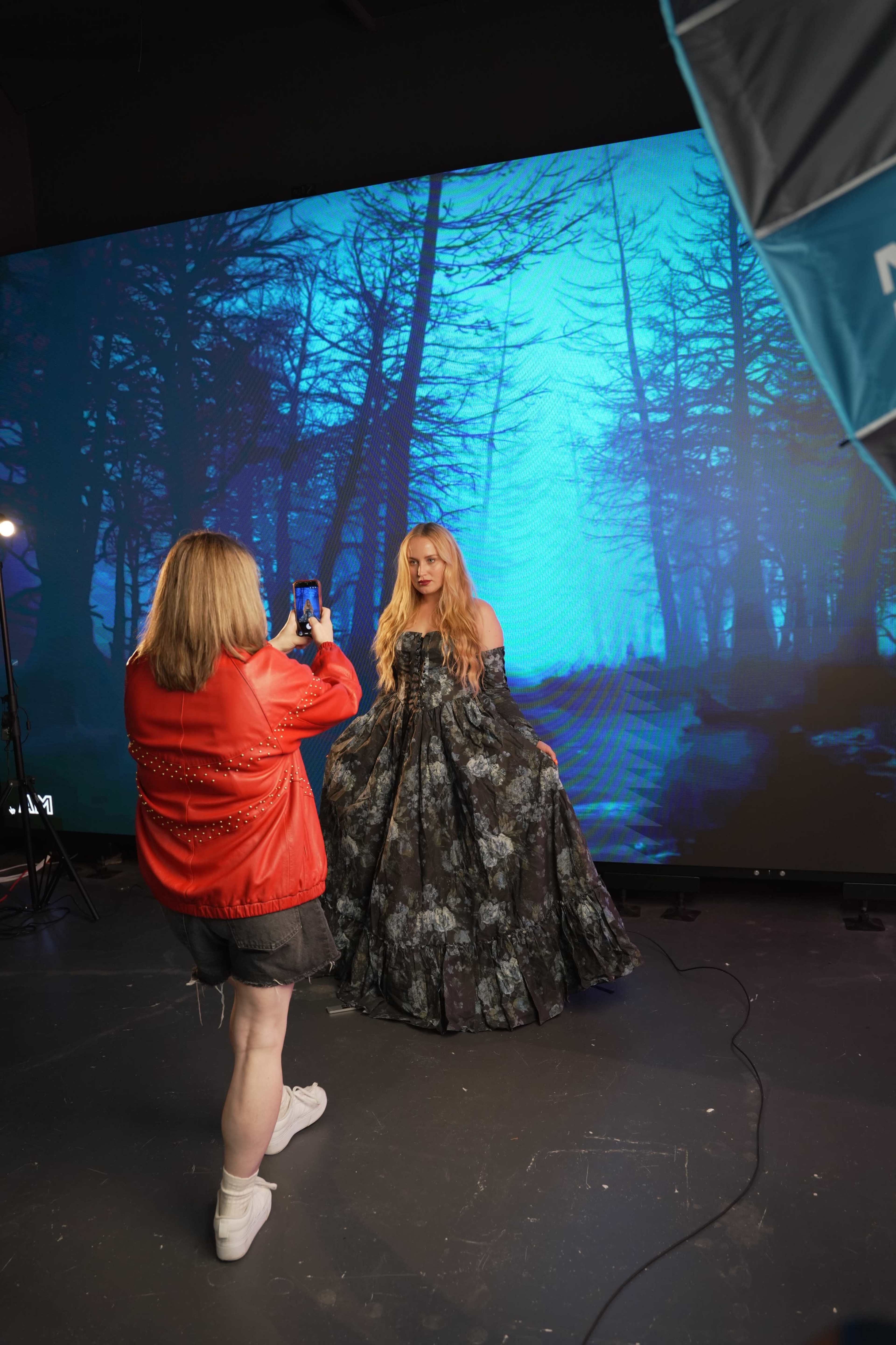 A woman in a red jacket takes a photo of a model wearing a dark floral gown in front of a large digital backdrop depicting a forest.