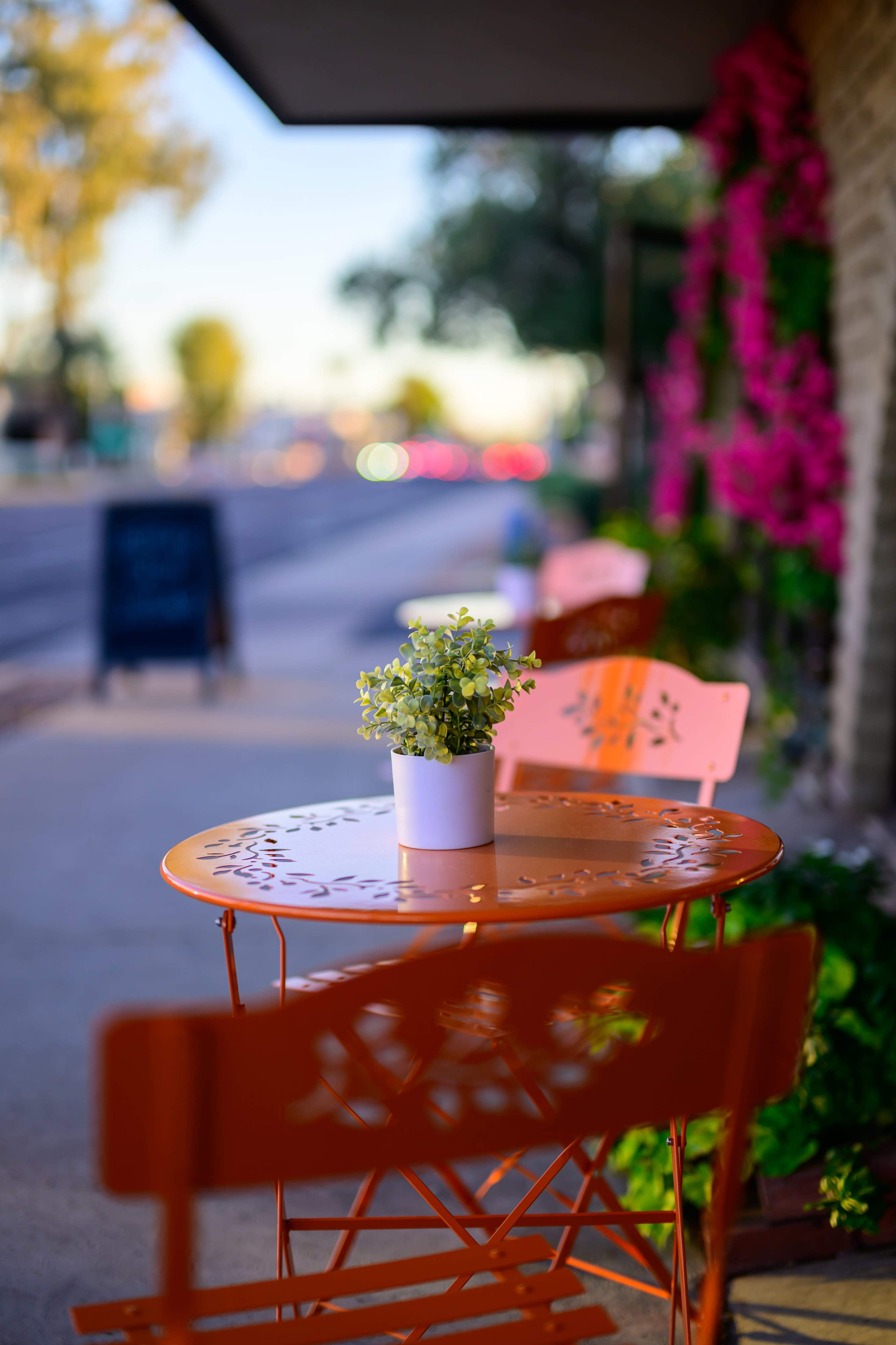 A small orange table with a white pot of greenery is set on a sidewalk beside a street lined with flowering plants.