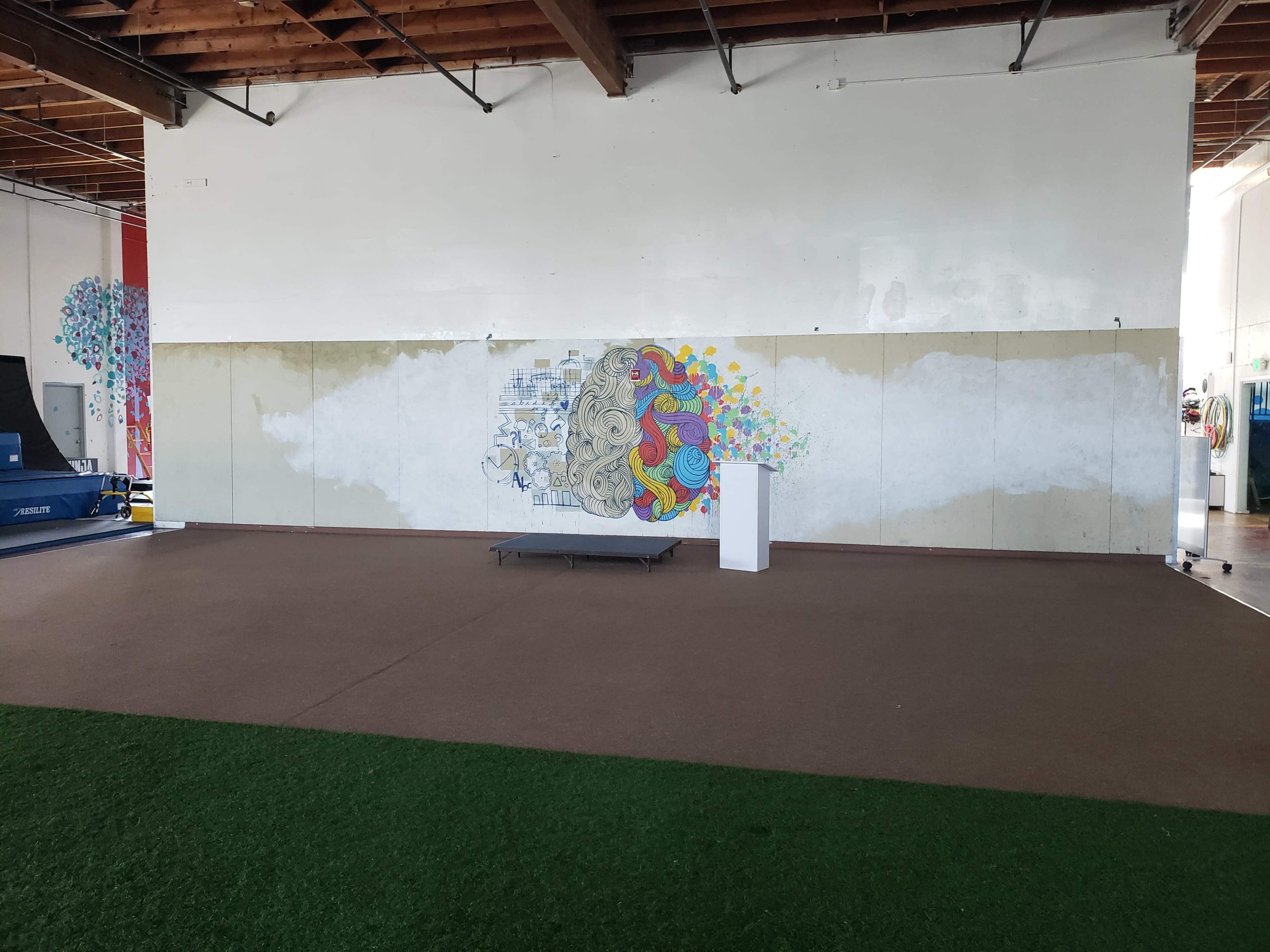 A large, partially painted wall featuring a colorful brain illustration, with a green carpeted area and a white pedestal in an unfinished indoor space.
