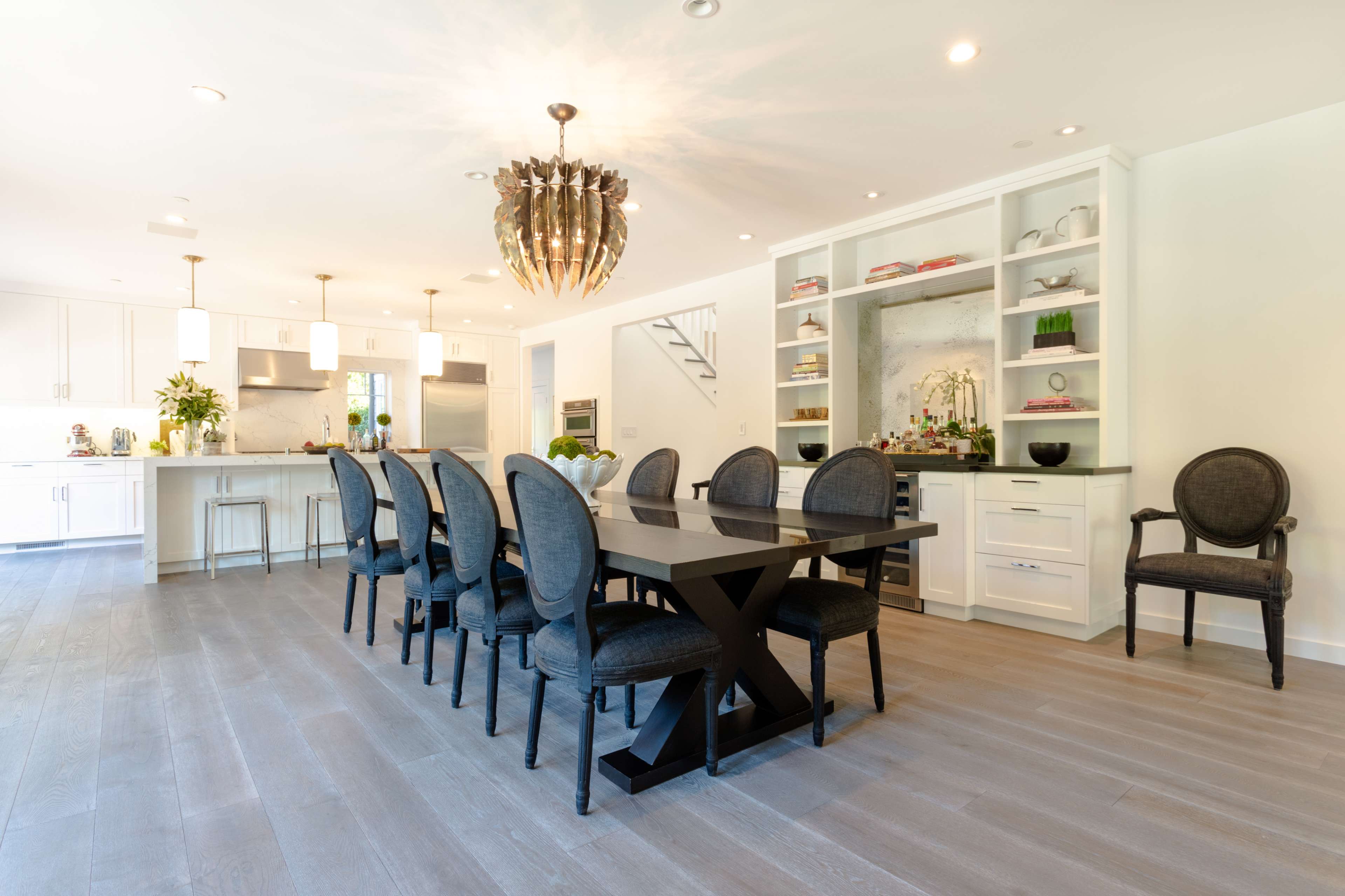 A modern kitchen and dining area featuring a long black dining table with eight chairs, a chandelier overhead, and white cabinetry with shelves displaying decorative items.