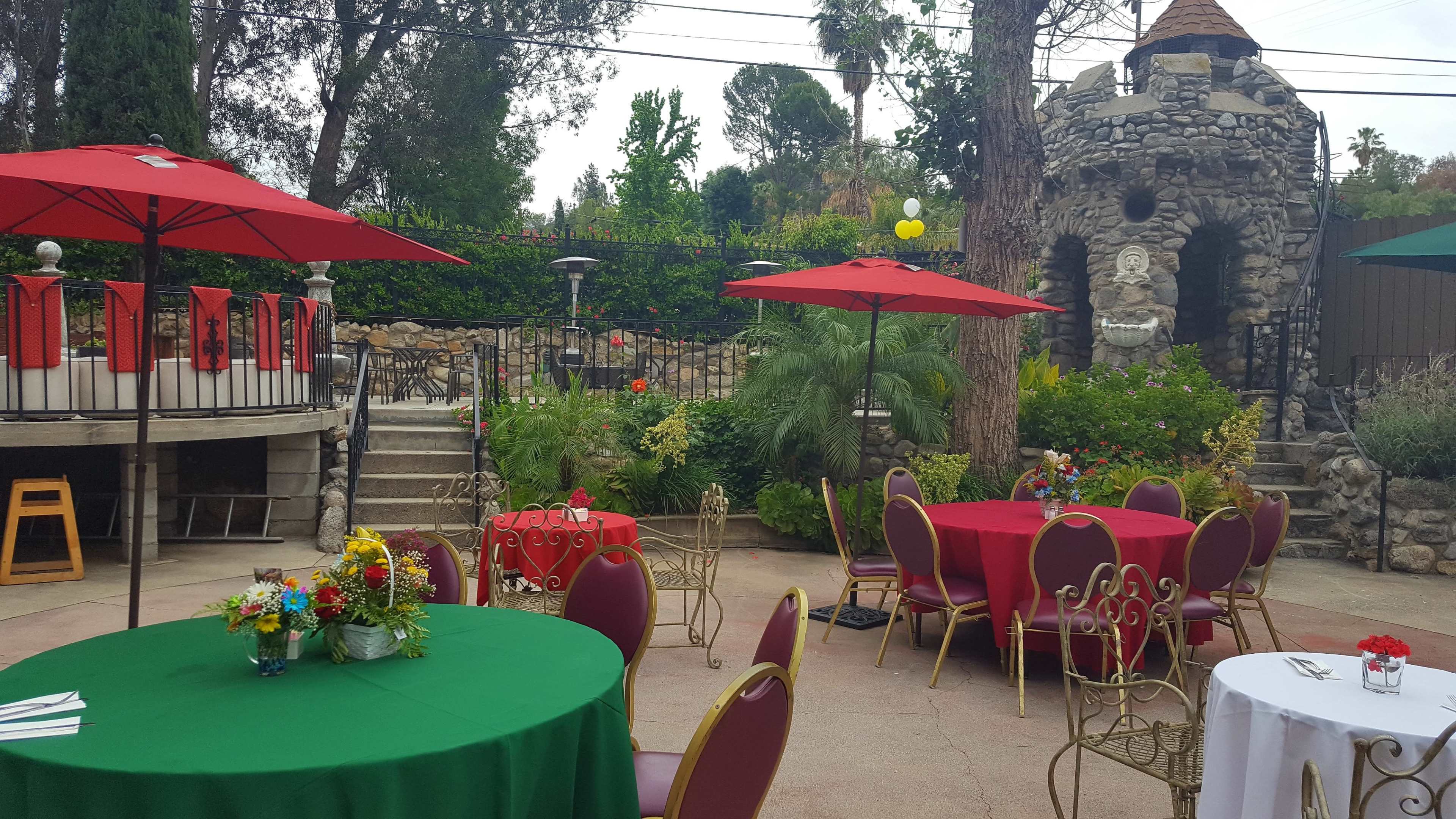 The outdoor dining area features circular tables covered in green and red cloths, surrounded by chairs, with umbrellas providing shade and a stone structure in the background.