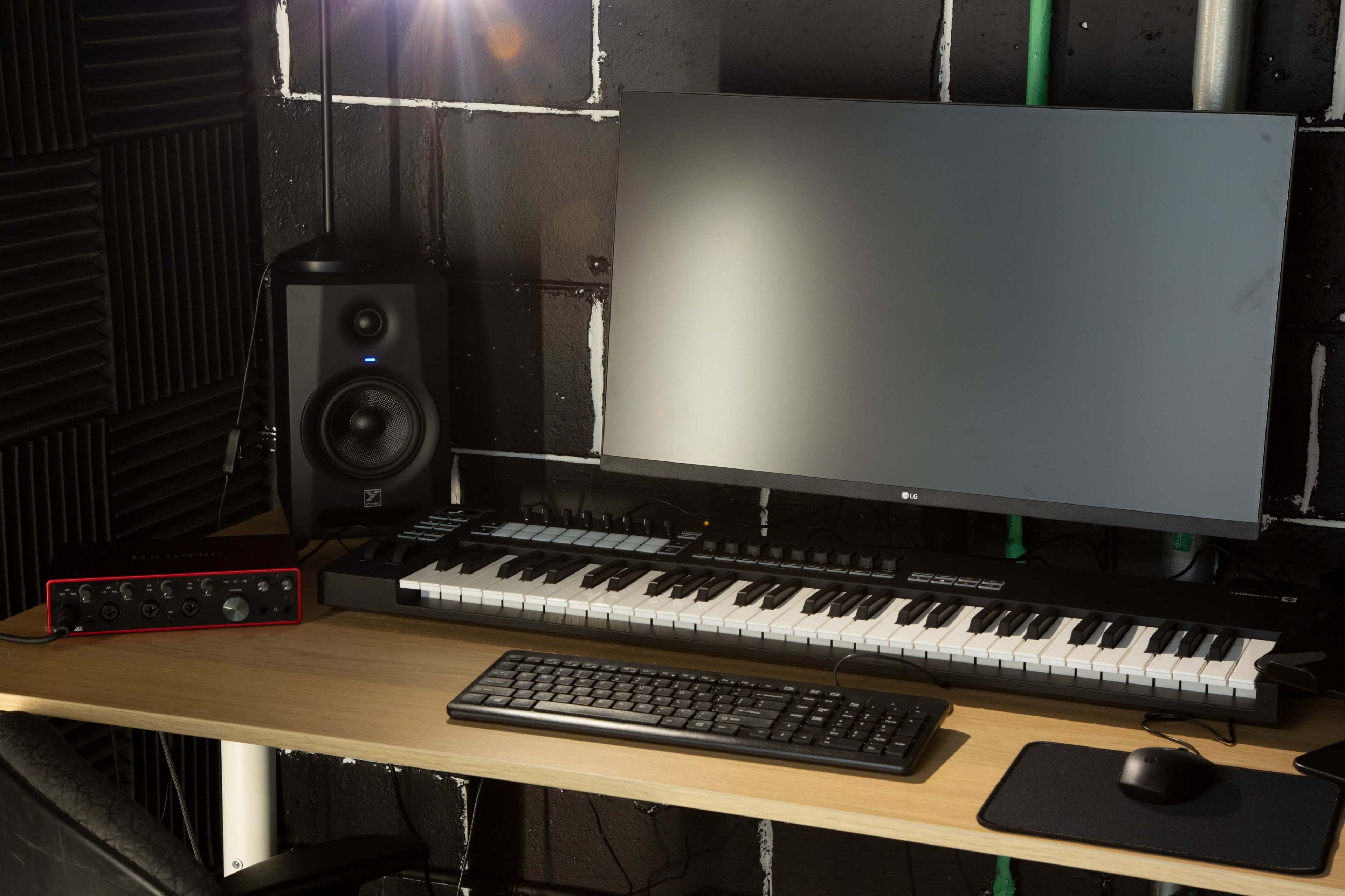 A desk setup features a computer monitor, MIDI keyboard, audio interface, and studio monitor speaker against a textured black wall.