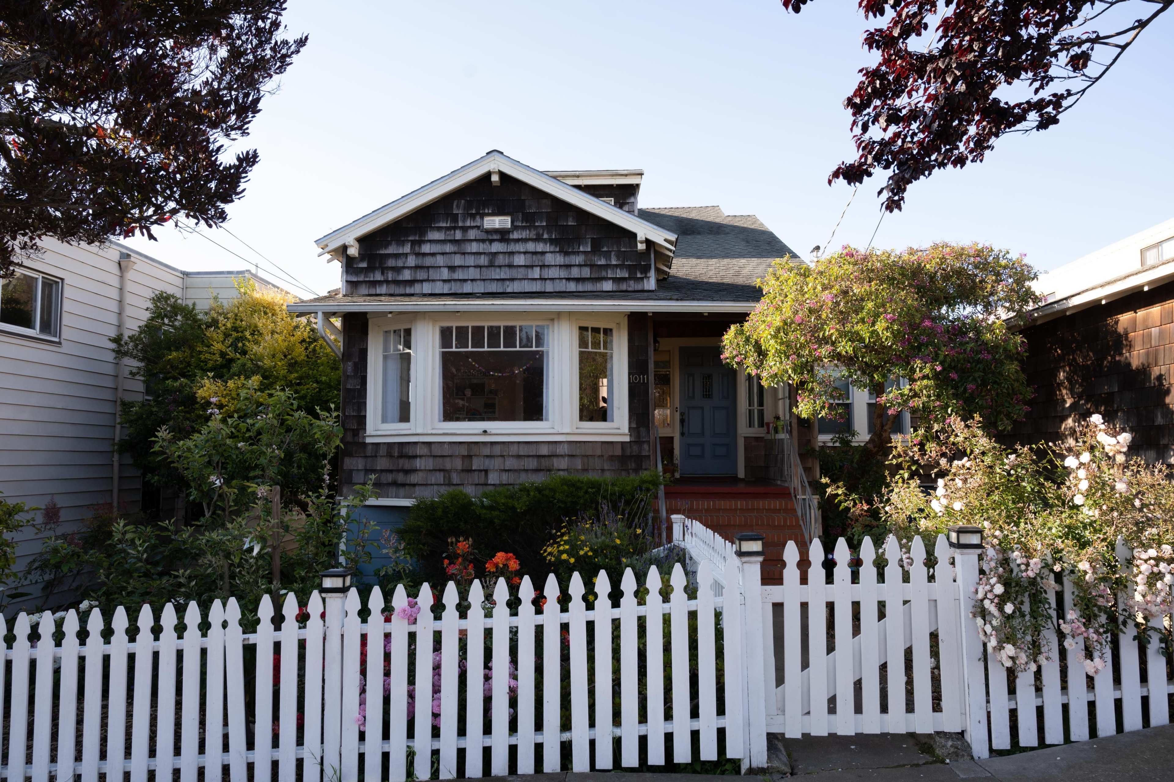 A wooden house with a shingled exterior and a front garden is enclosed by a white picket fence.