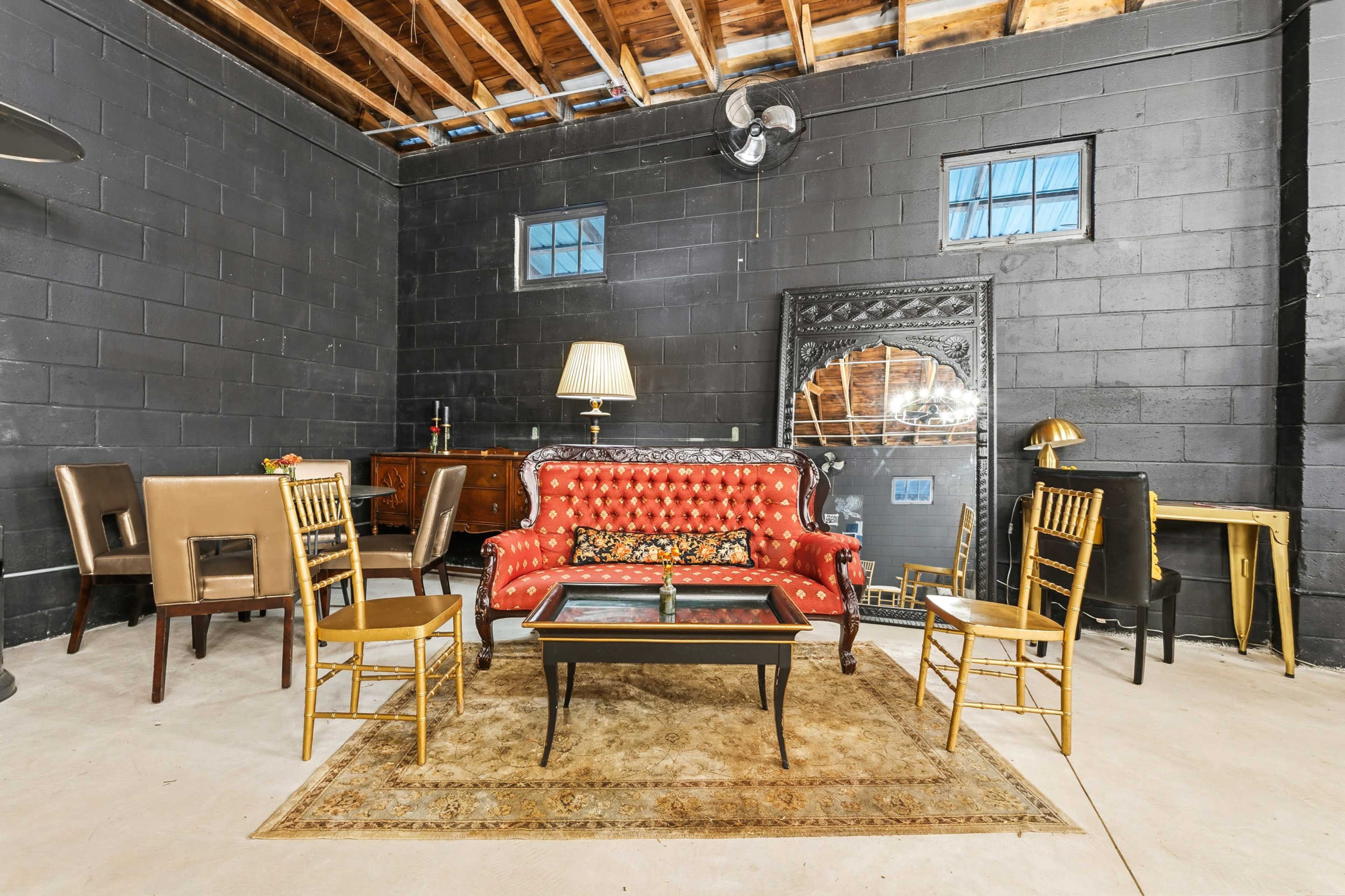 The scene features a vintage-style sitting area with a red tufted sofa, a glass coffee table, and gold chairs arranged around an area rug in a space with black walls and exposed wooden beams.