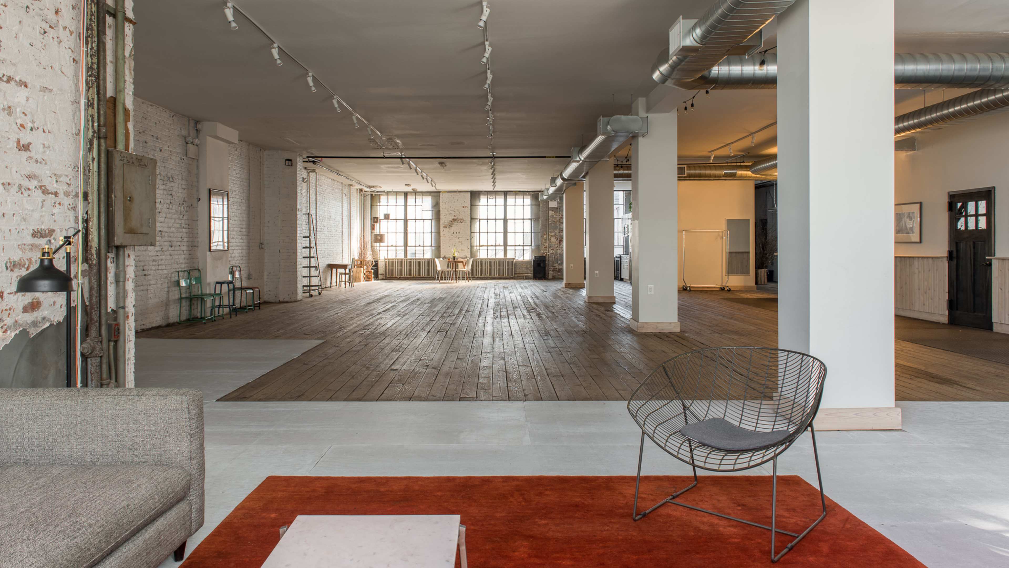 The image shows a spacious, modern loft with large windows, hardwood floors, and minimal furnishings.
