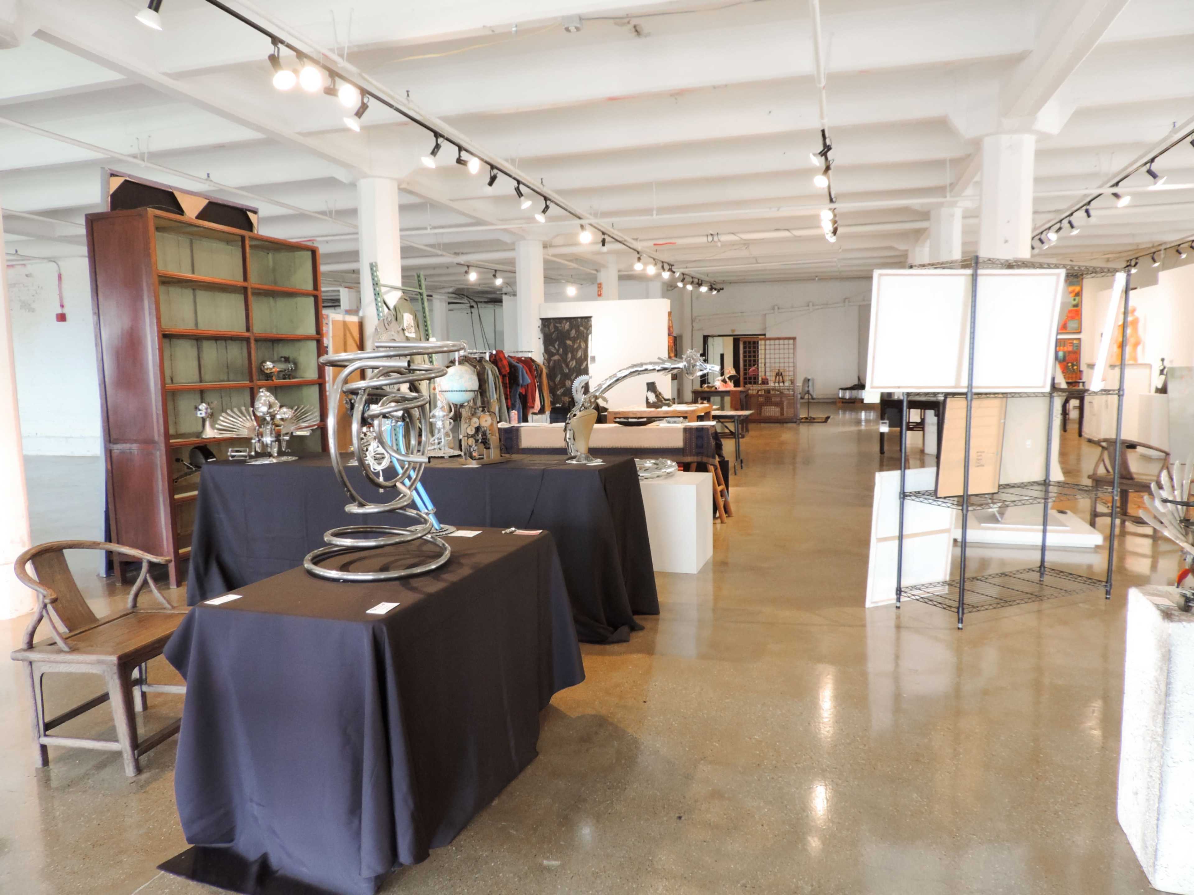 The image shows an art gallery with various sculptures and furniture displayed on black tables in a spacious, well-lit room.